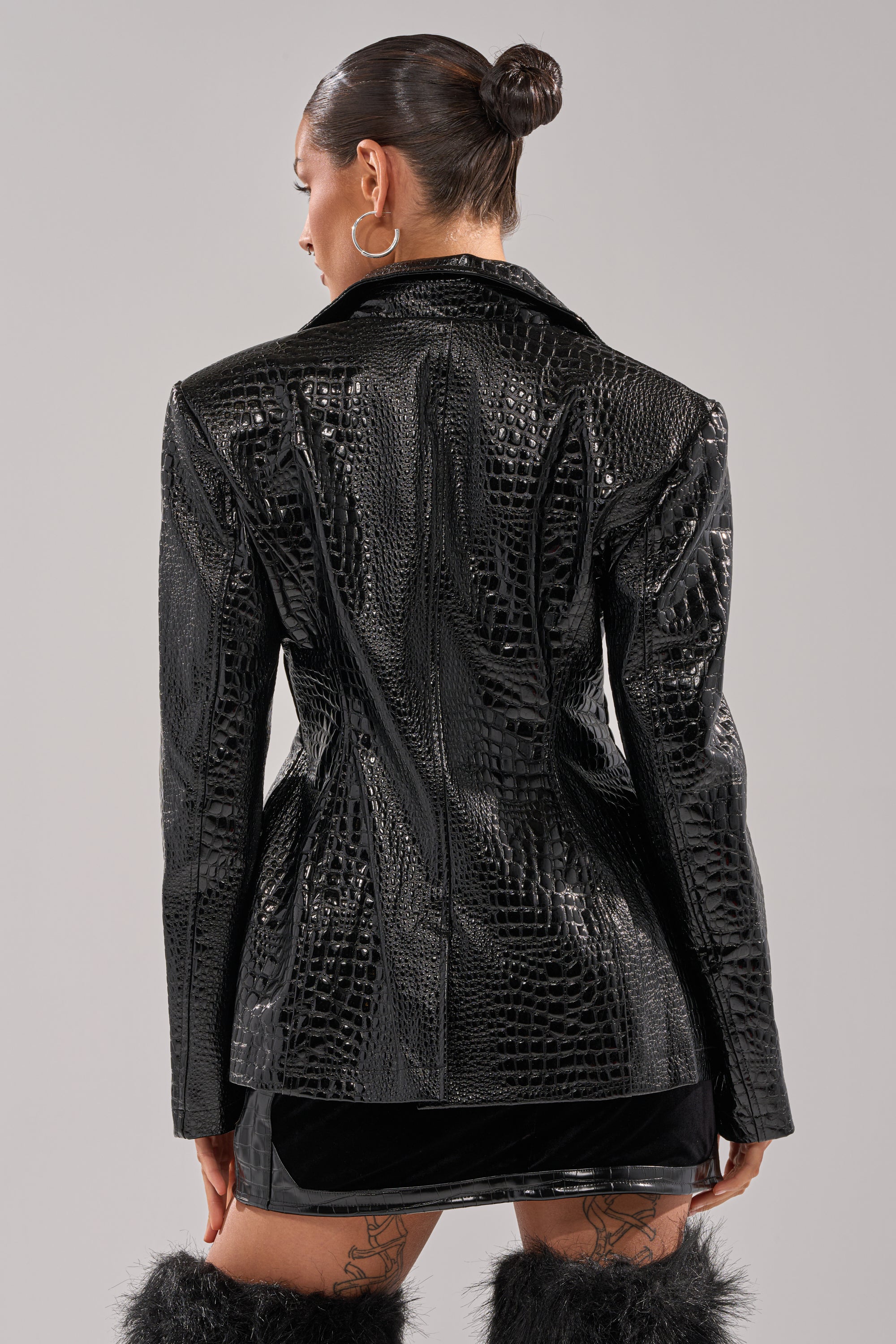 A woman with her hair in a bun wears the FOR THE DRAMA CROC BLAZER, a faux patent leather jacket with a crocodile print, paired with a short black skirt and black furry boots. Facing away, she shows off hoop earrings and leg tattoos.