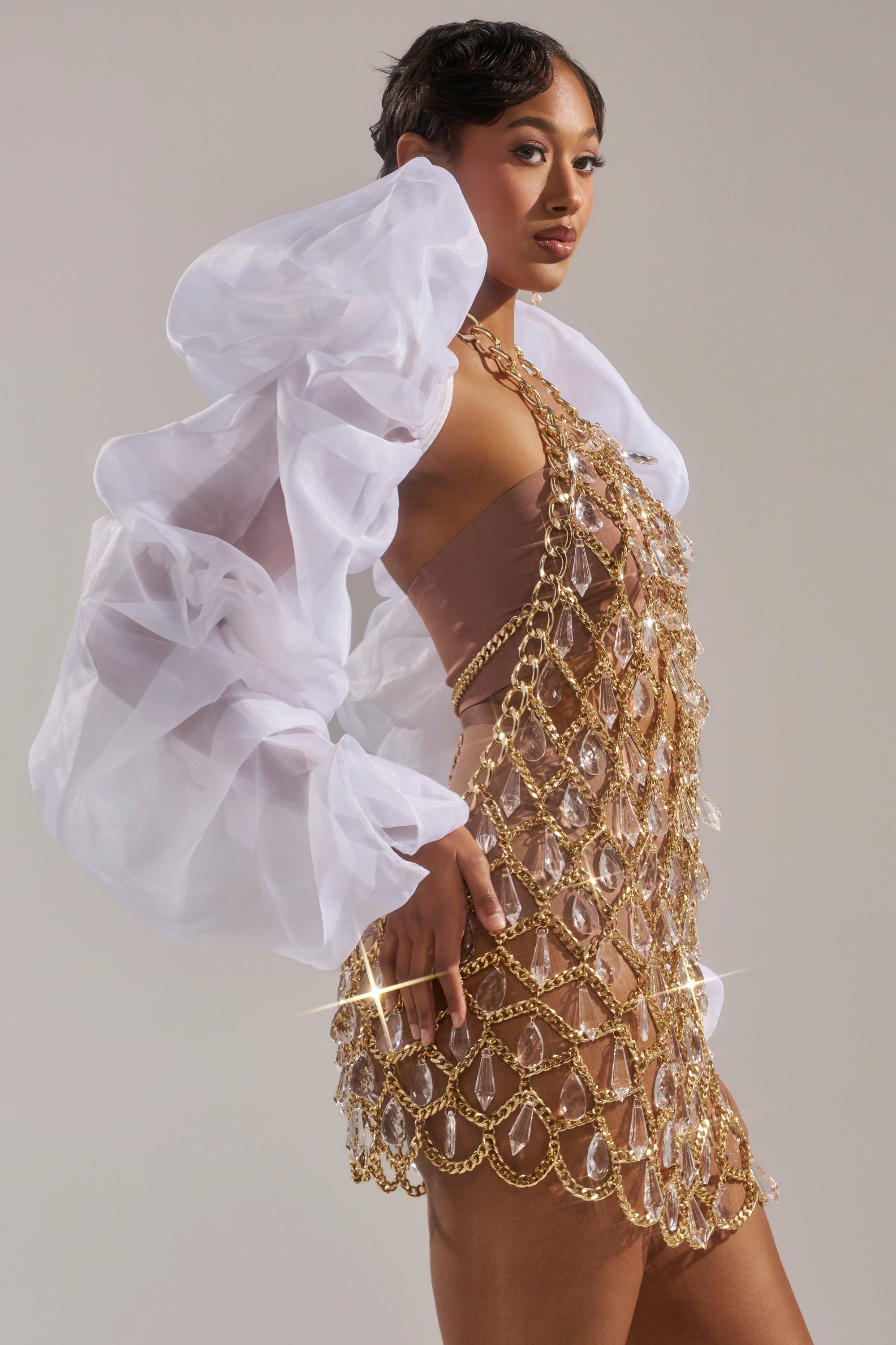 A woman poses confidently against a neutral background, wearing the MAIN SQUEEZE ORGANZA SHRUG—sheer white puff sleeves paired with a nude-toned mini dress adorned with gold chains and clear crystal embellishments.