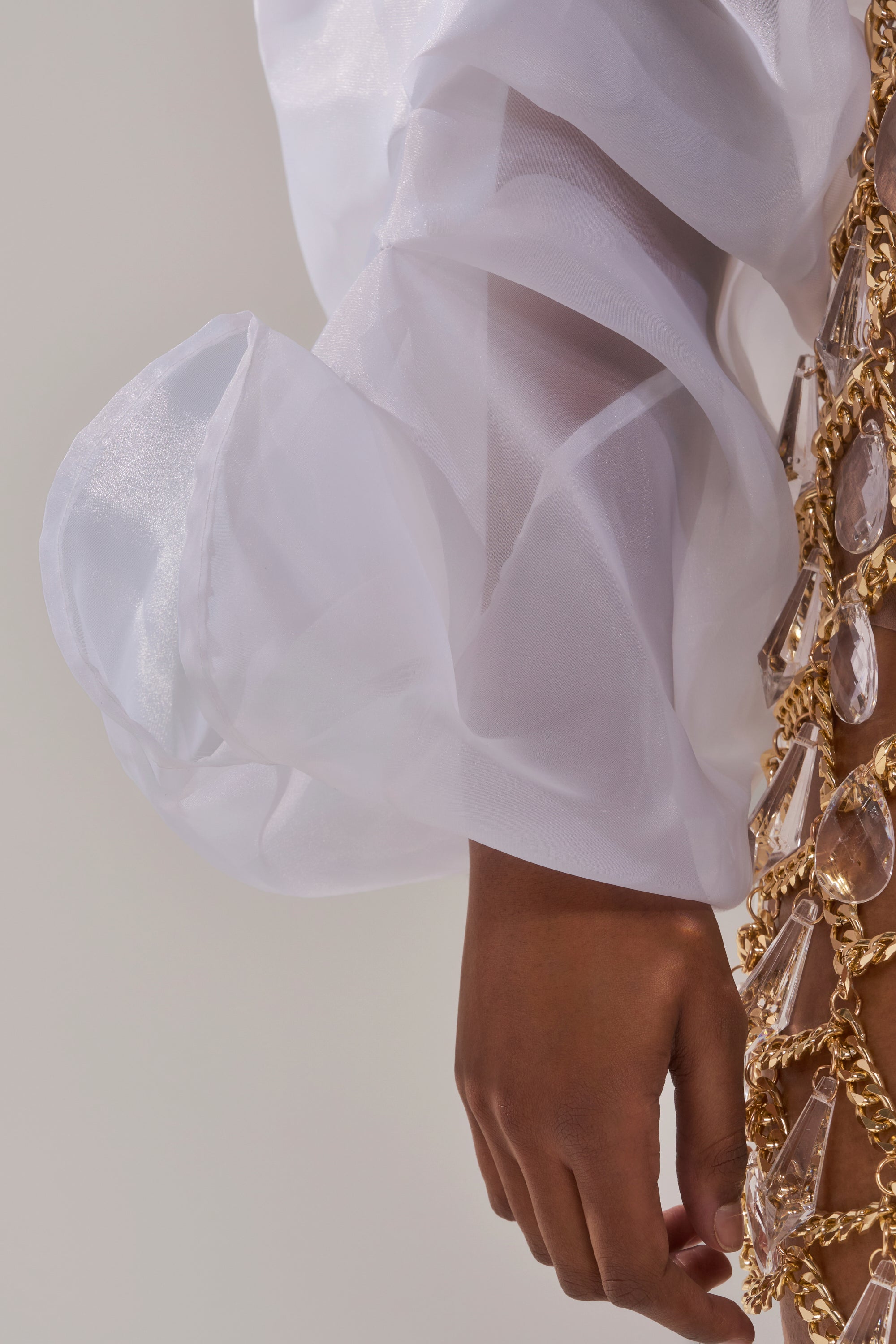 A close-up of a hand wearing the MAIN SQUEEZE ORGANZA SHRUG with sheer white puffy sleeves, holding a gold chain adorned with large clear gemstones against a plain, neutral background.