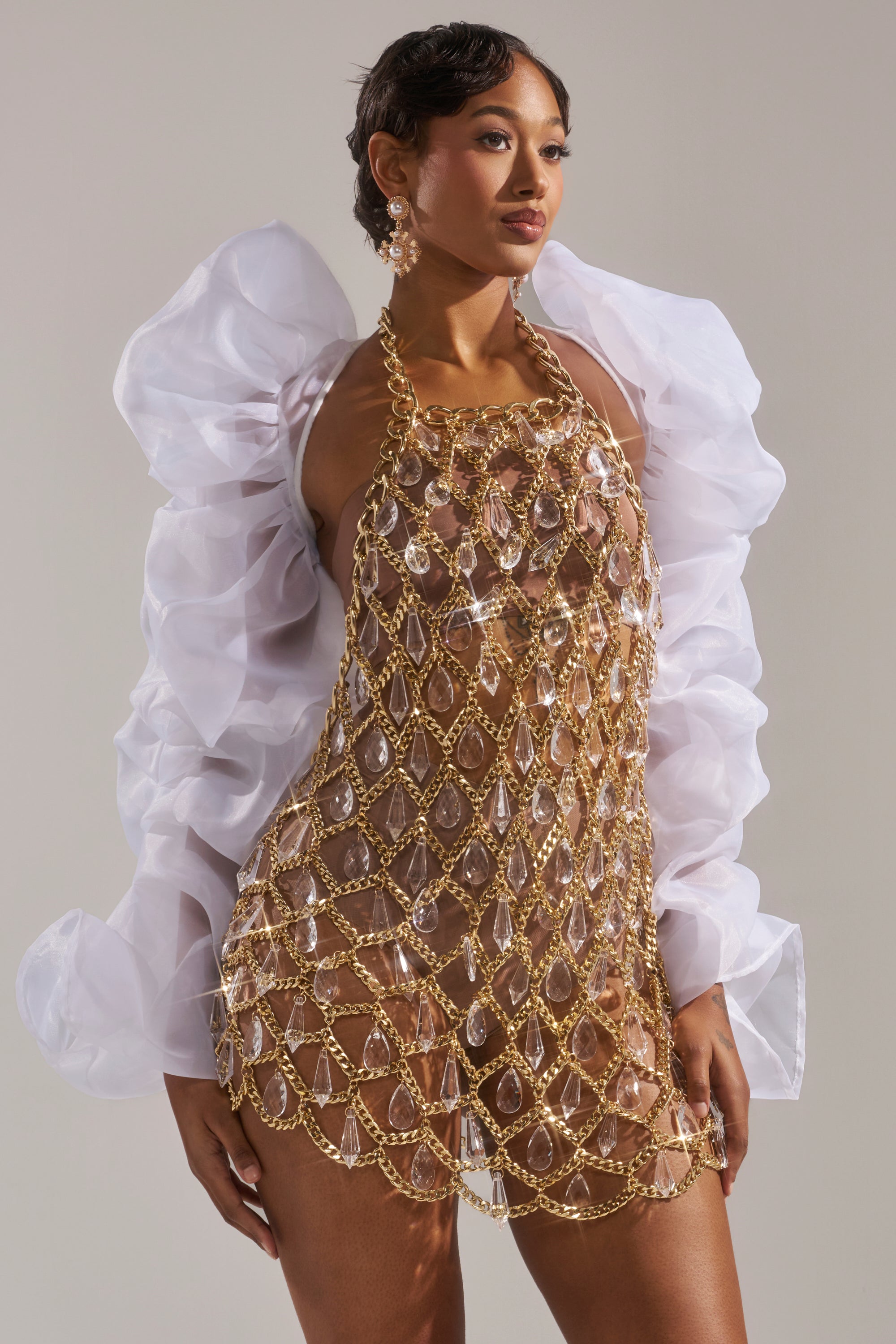 A woman poses in the MAIN SQUEEZE ORGANZA SHRUG with puff sleeves, styled over a gold chain and crystal mesh dress, nude bodysuit, and gold earrings, set against a plain light background.