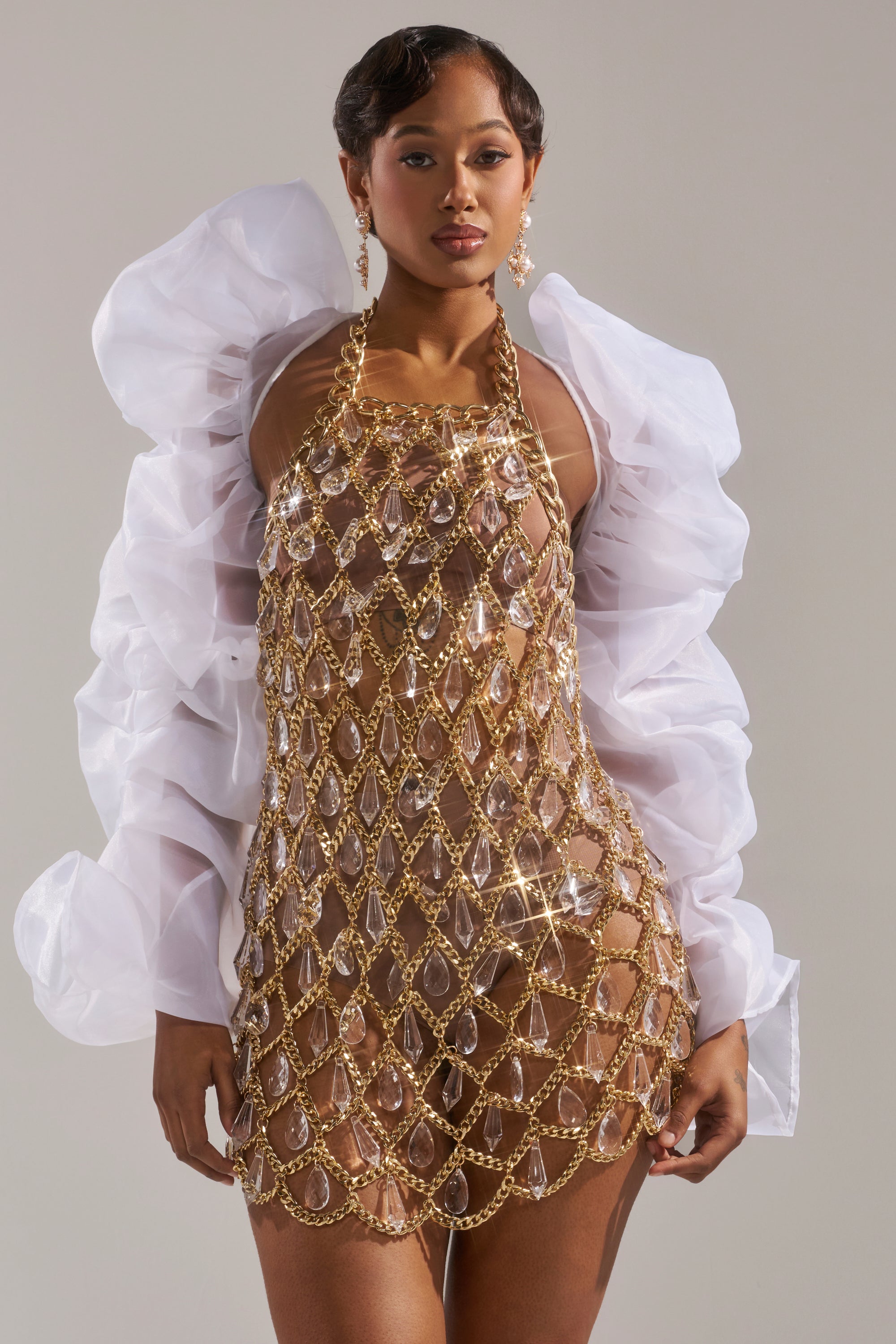 A woman poses confidently in the MAIN SQUEEZE ORGANZA SHRUG, featuring voluminous puff sleeves over a sheer gold chain dress with crystal-like gems and a nude bodysuit. Matching earrings complete her striking cropped silhouette against a plain background.