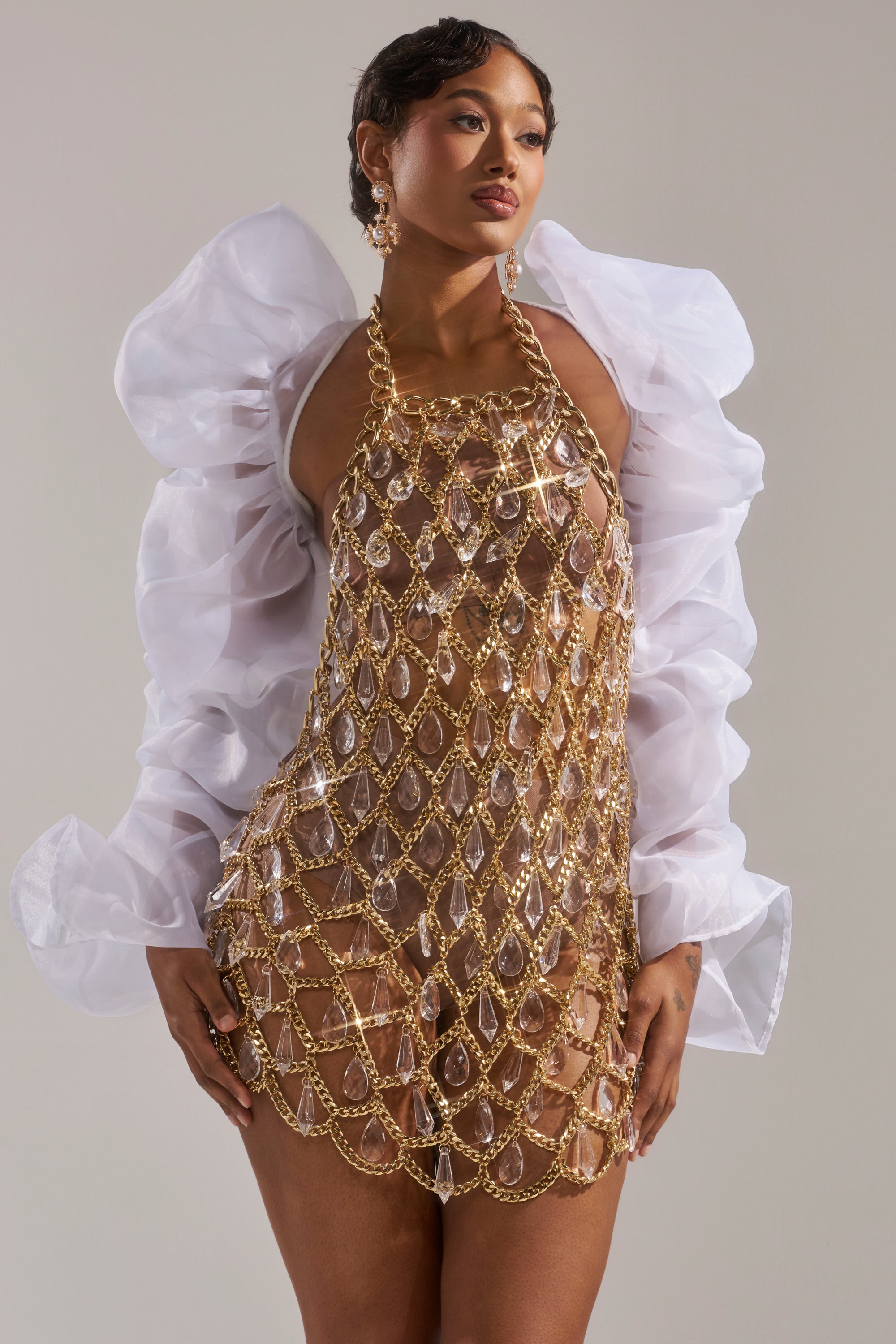 A woman models the MAIN SQUEEZE ORGANZA SHRUG with puff sleeves over a sheer, gold chain and crystal mesh dress and nude bodysuit, accessorized with statement earrings. She stands against a plain background, her short hair styled sleekly.