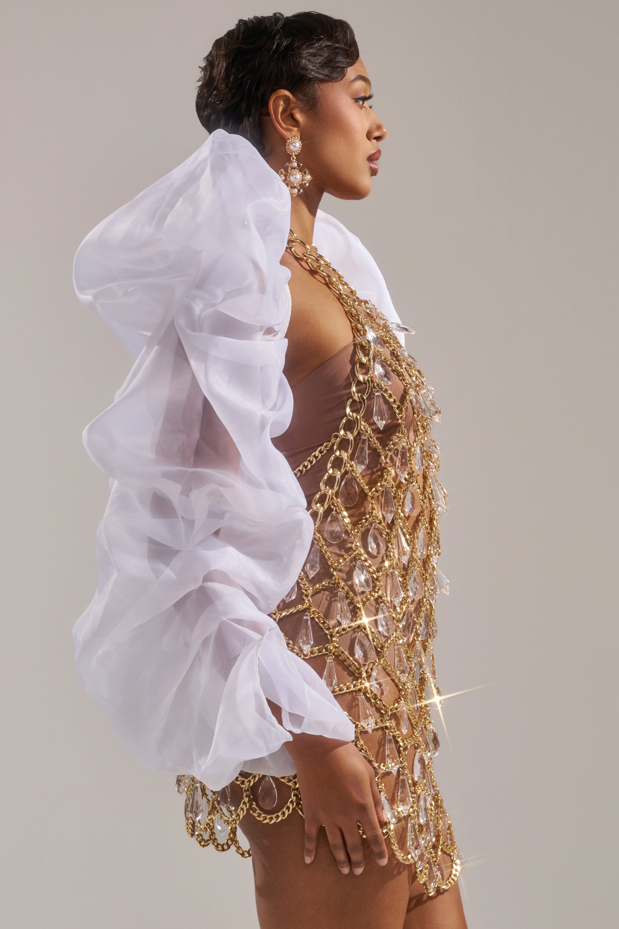 A woman poses in profile against a plain light background, wearing the MAIN SQUEEZE ORGANZA SHRUG with white ruffled sleeves over a sheer gold chain and crystal dress layered on a nude bodysuit, styled with large earrings.