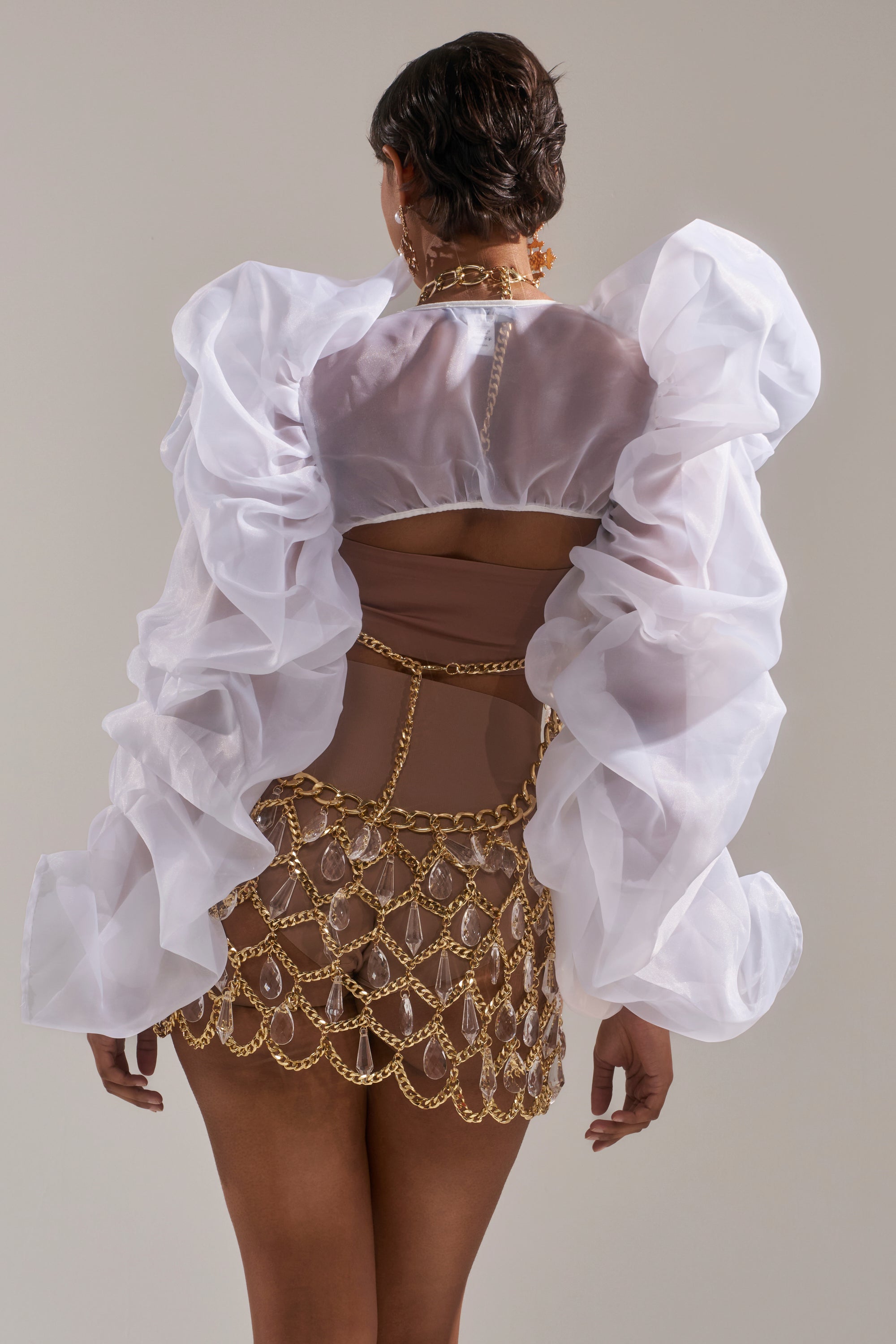 A person stands facing away, wearing the MAIN SQUEEZE ORGANZA SHRUG featuring sheer white fabric and dramatic ruffled sleeves, paired with a gold chain skirt over a nude bodysuit against a neutral background.