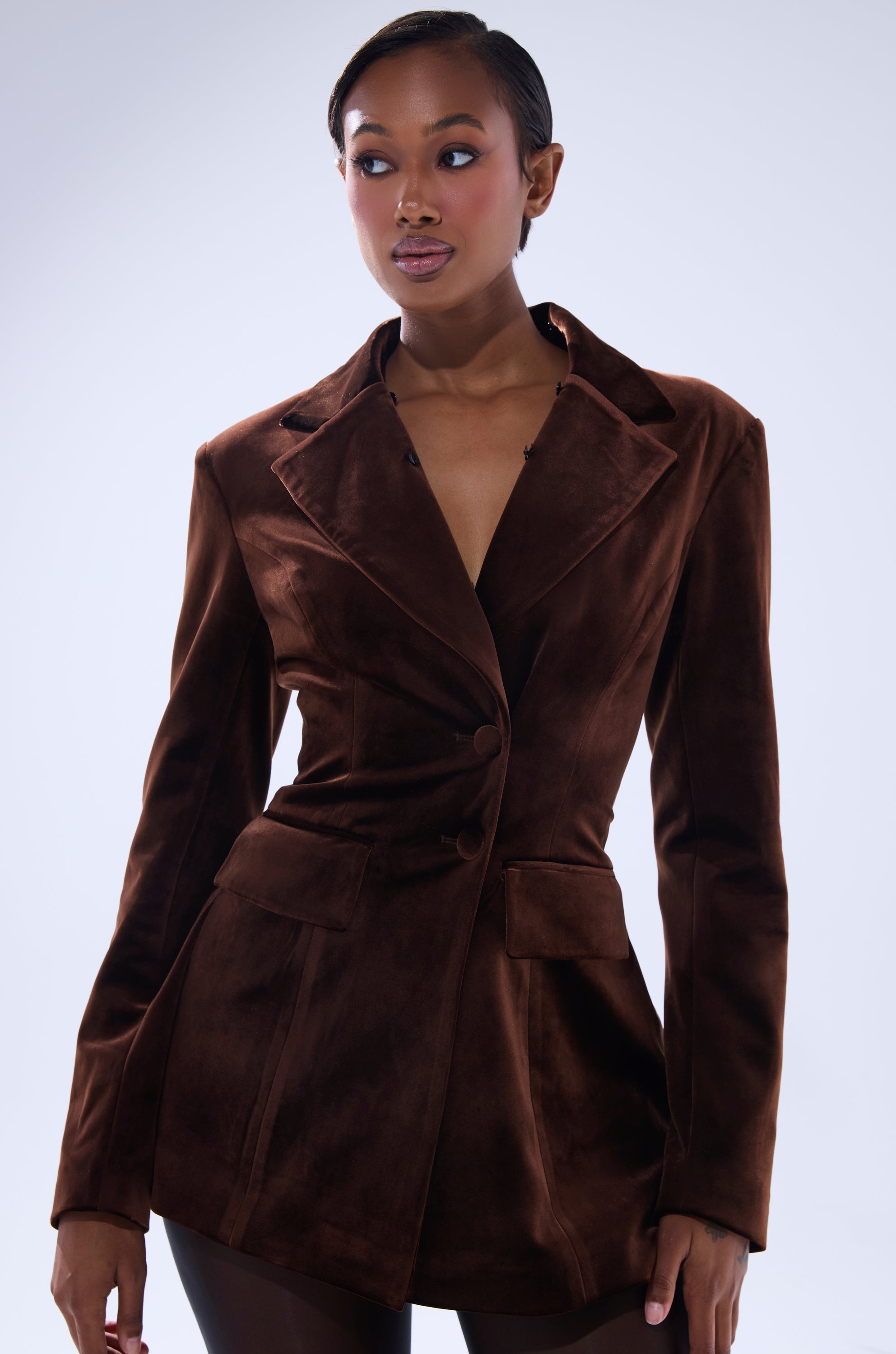 A woman stands against a light background, confidently gazing to the side in the fitted SUNDAY VELVET BLAZER WITH REMOVABLE FUR COLLAR, featuring large lapels and front pockets.