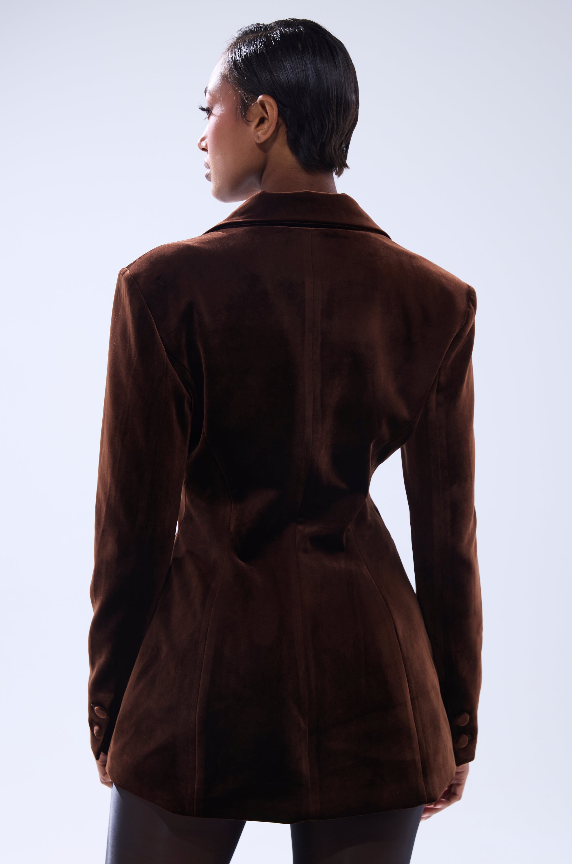 A person with short, slicked-back hair is seen from behind wearing the SUNDAY VELVET BLAZER WITH REMOVABLE FUR COLLAR in dark brown and black leggings, set against a plain light background.