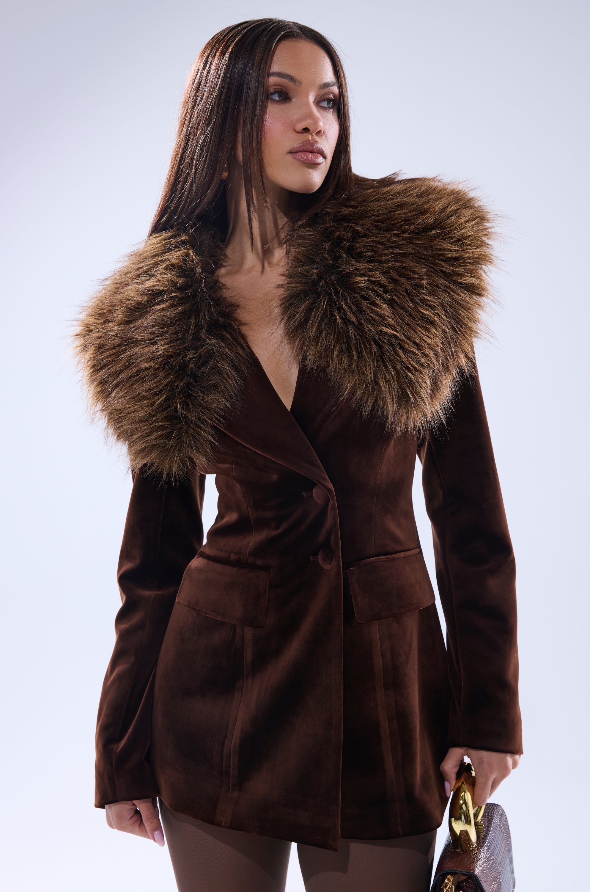 A woman stands confidently against a light background, holding a small handbag and wearing the SUNDAY VELVET BLAZER WITH REMOVABLE FUR COLLAR. She has long straight hair and gazes slightly to the side.