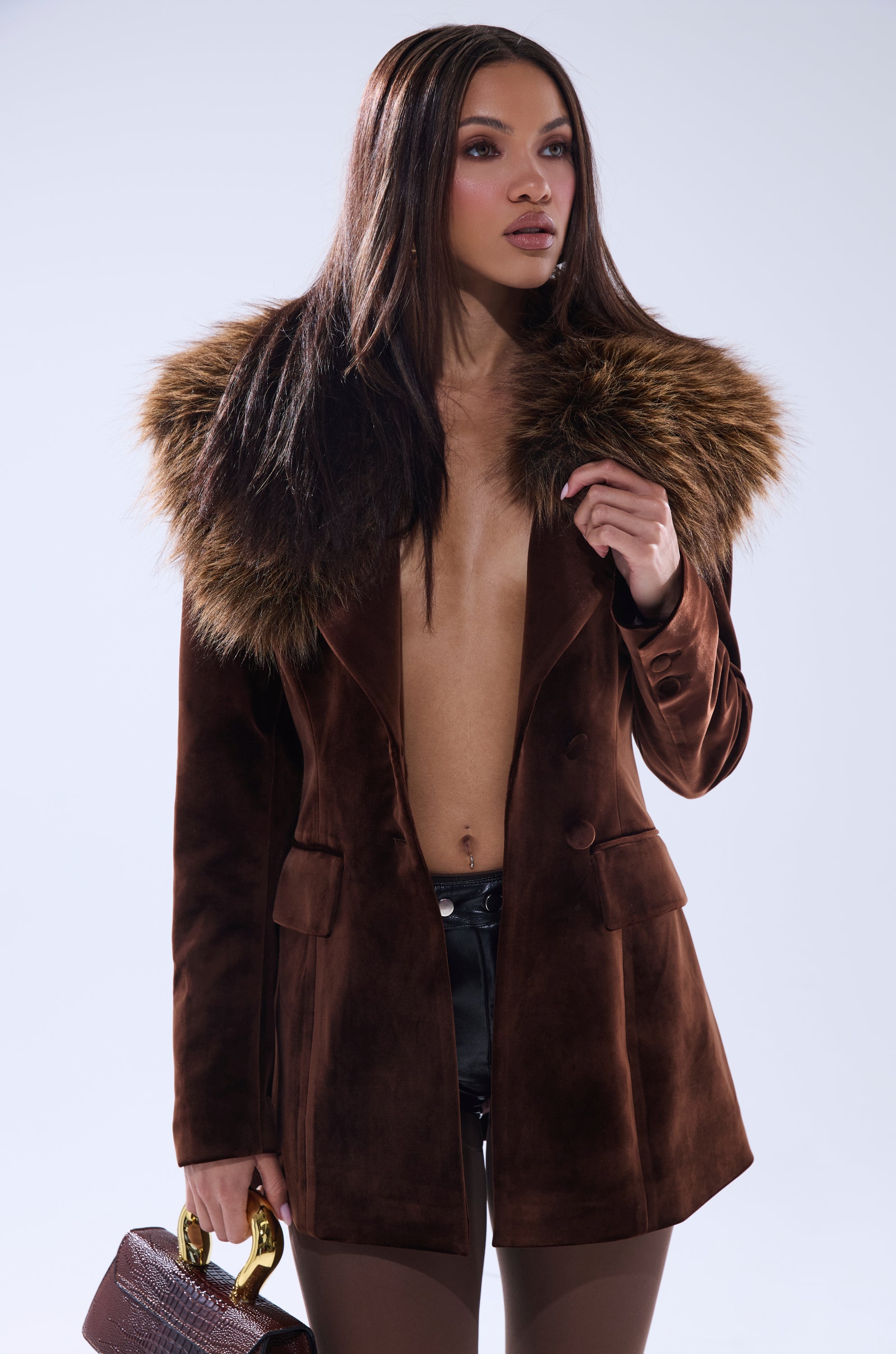 A woman models the SUNDAY VELVET BLAZER WITH REMOVABLE FUR COLLAR in brown, partially open to reveal her chest. She holds a textured brown handbag and poses with straight, long hair against a plain light background.