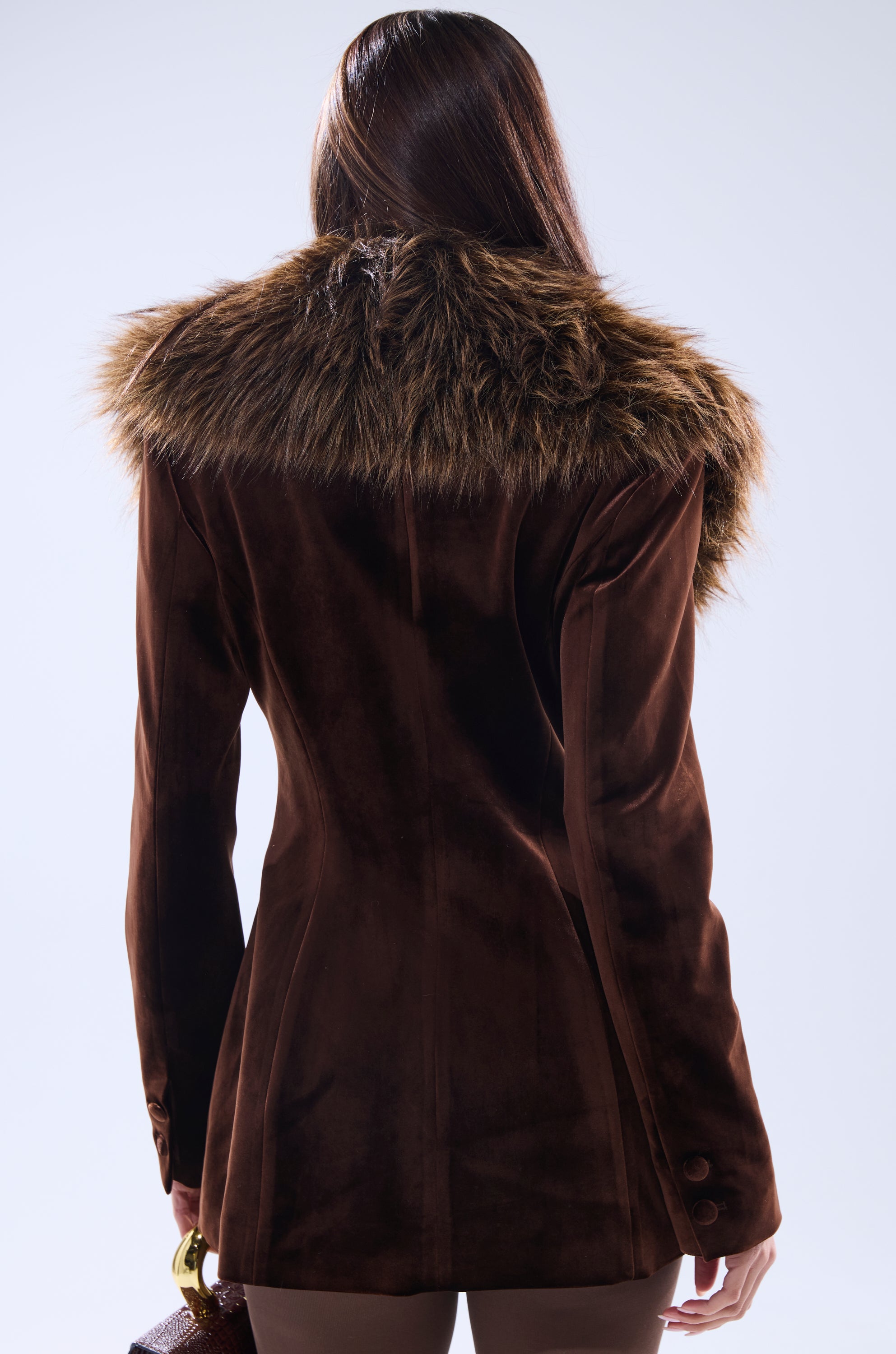 A person with long, straight brown hair is seen from behind wearing the SUNDAY VELVET BLAZER WITH REMOVABLE FUR COLLAR. The background is plain and light, and they are holding a small purse.
