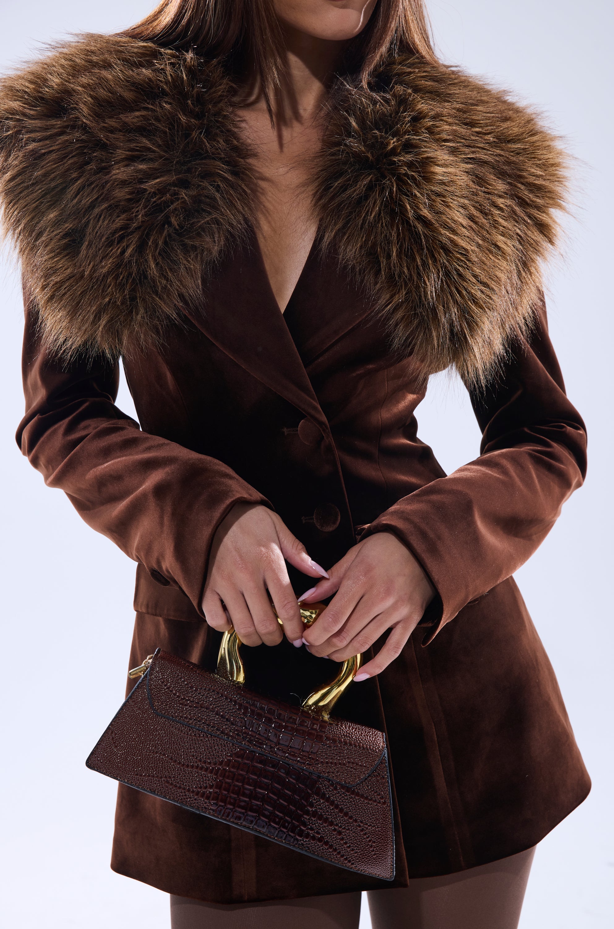 A woman wears the SUNDAY VELVET BLAZER WITH REMOVABLE FUR COLLAR, holding a dark brown textured handbag with gold handles. Her face is partially out of frame as she stands against a light background.