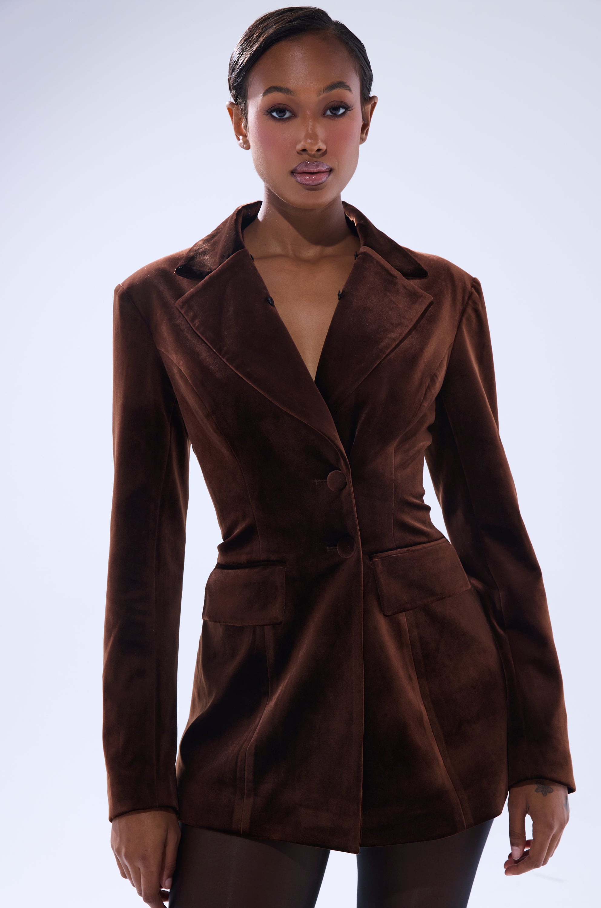 A woman stands confidently in the SUNDAY VELVET BLAZER WITH REMOVABLE FUR COLLAR, tailored and buttoned at the waist with two front pockets. She has short styled hair and natural makeup, posing against a light background.