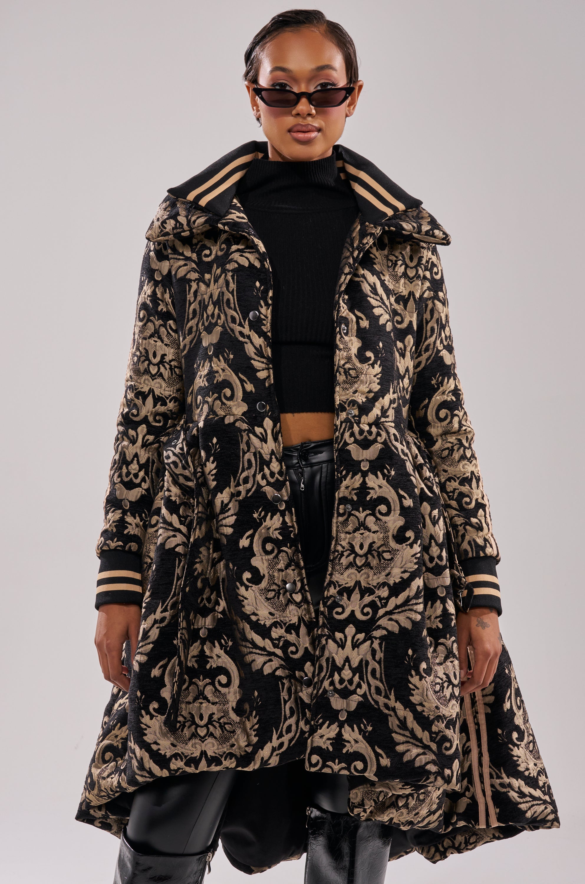 A woman in dark sunglasses, a black crop top, and shiny black pants stands confidently in the DECKER PUFFER TRENCH IN JACQUARD, featuring an ornate black and gold pattern with a wide collar, striped trim, and belted waist.