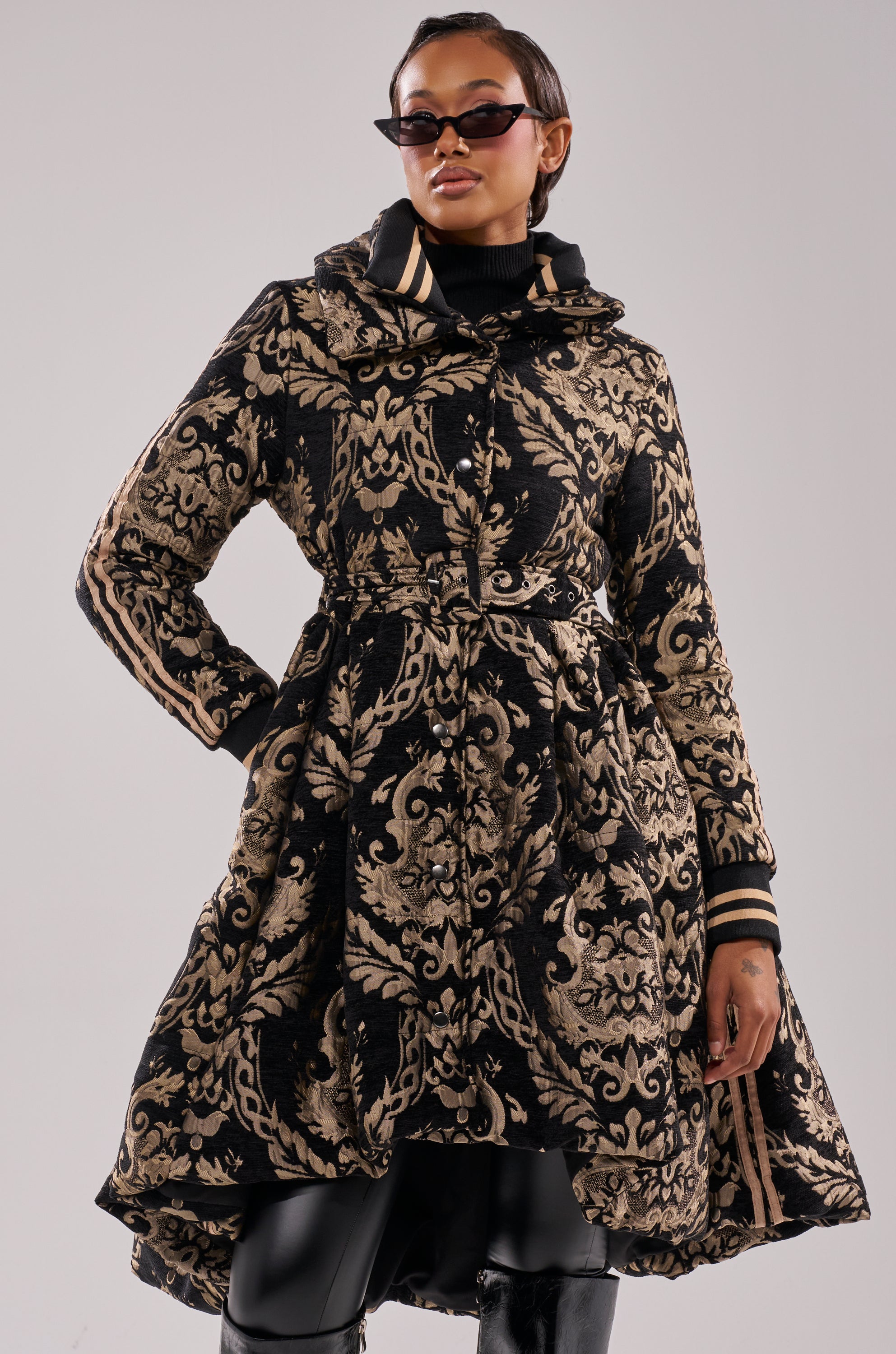 Wearing the DECKER PUFFER TRENCH IN JACQUARD, a black and gold jacket with a flared hem, paired with a black turtleneck, sunglasses, and pants, a person poses confidently against a plain light background.
