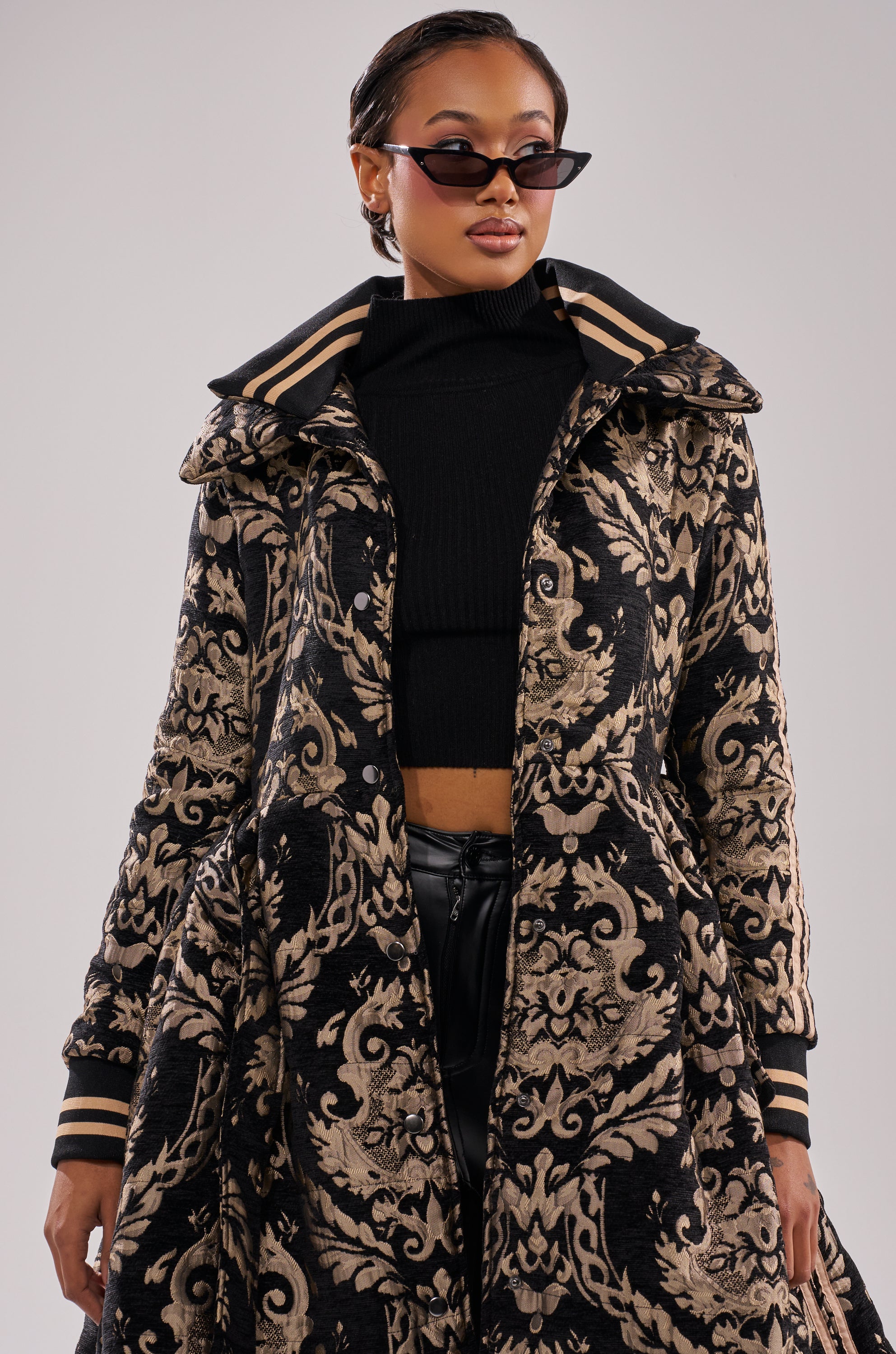 A woman stands confidently against a neutral background wearing cat-eye sunglasses, a black turtleneck crop top, and the DECKER PUFFER TRENCH IN JACQUARD.
