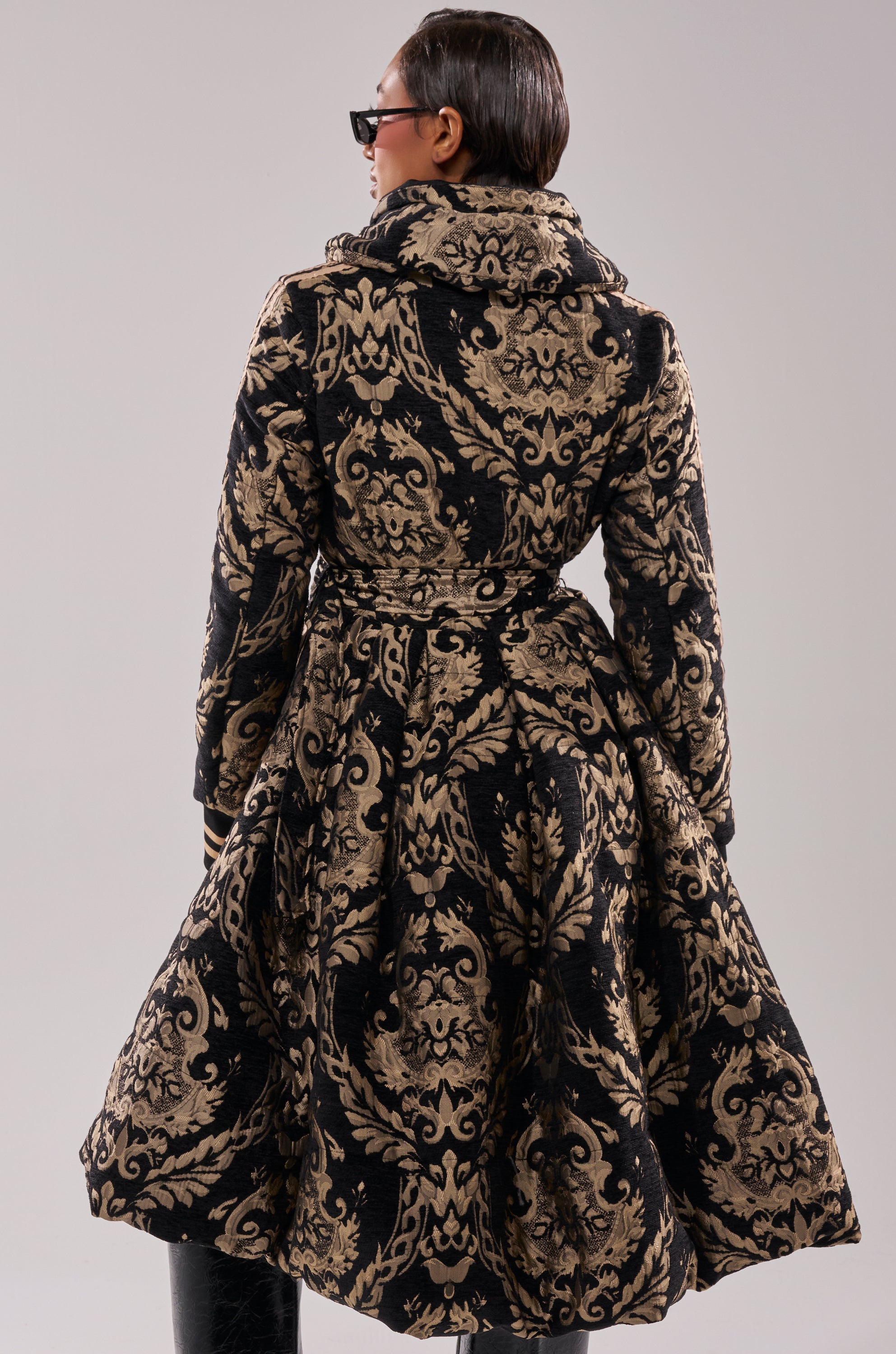 A person with short, slicked-back hair and sunglasses is seen from behind wearing the DECKER PUFFER TRENCH IN JACQUARD, featuring black and gold jacquard fabric and a full skirt, against a plain background.