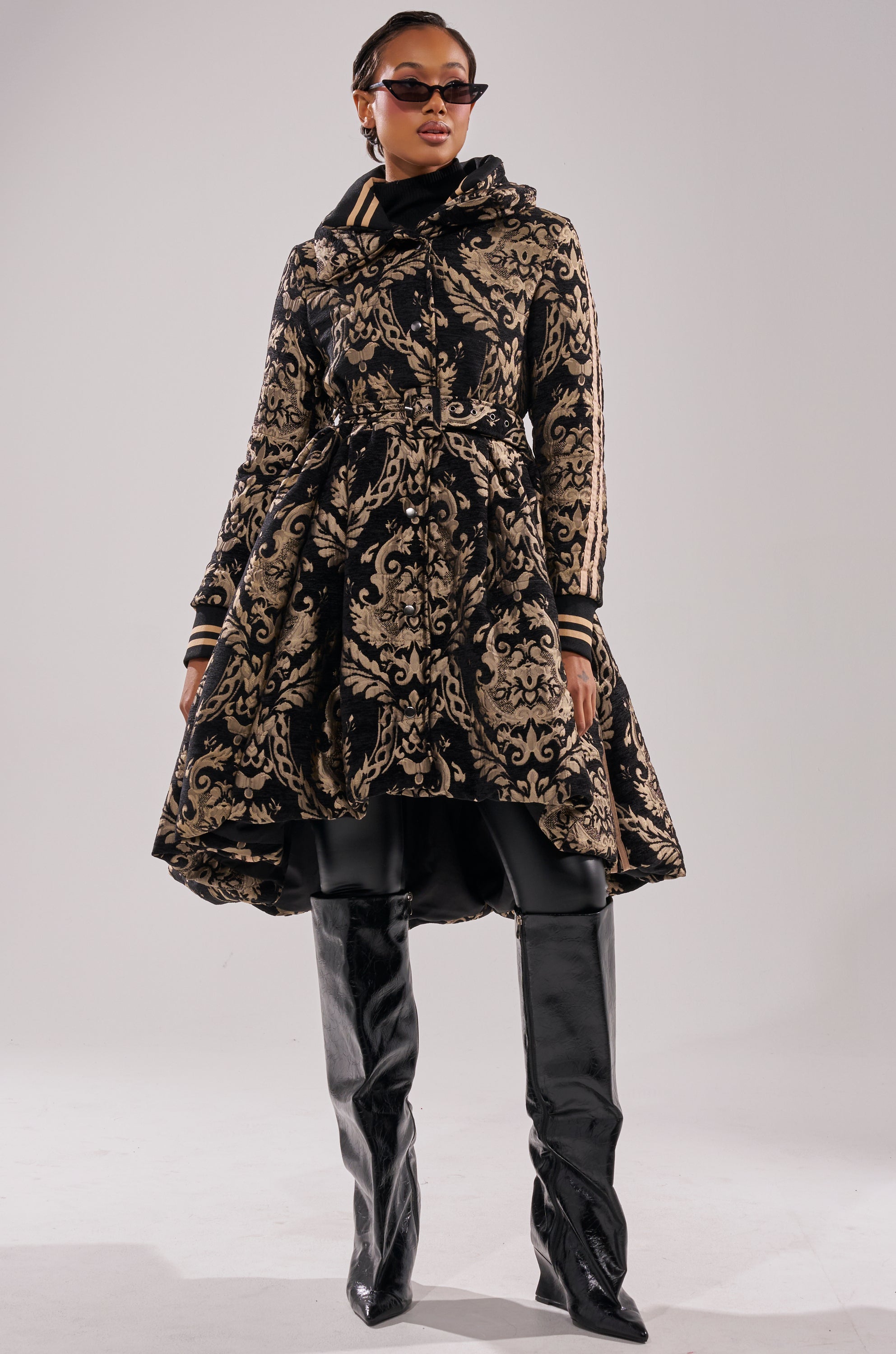 A woman in sunglasses and the DECKER PUFFER TRENCH IN JACQUARD, featuring a belted waist, black gloves, and shiny over-the-knee boots, stands confidently against a plain background.