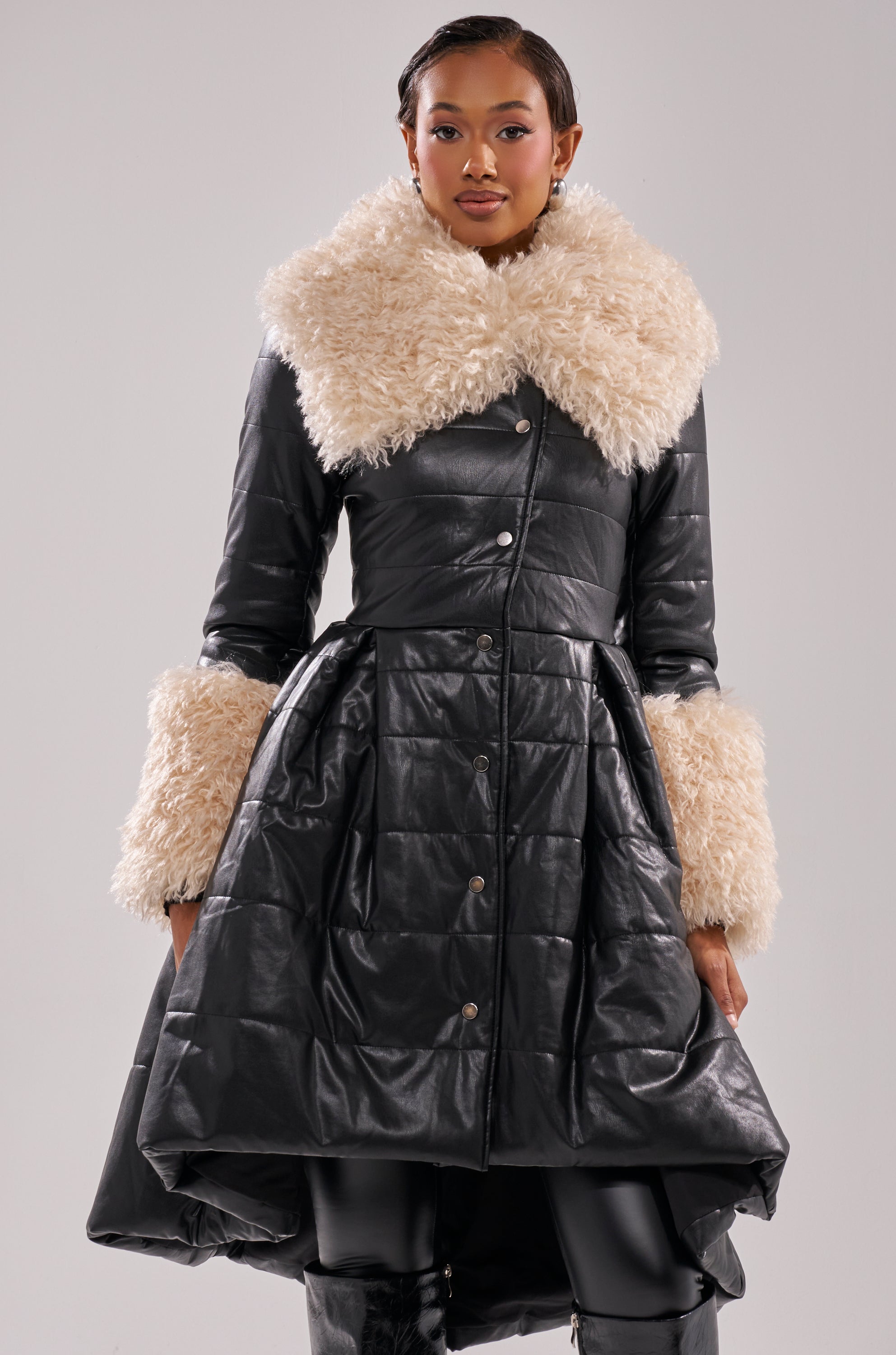 A woman stands confidently against a gray background, wearing the MIDTOWN PEPLUM PUFFER IN FAUX LEATHER—a black, fitted jacket with large cream faux fur trim on the collar and cuffs.