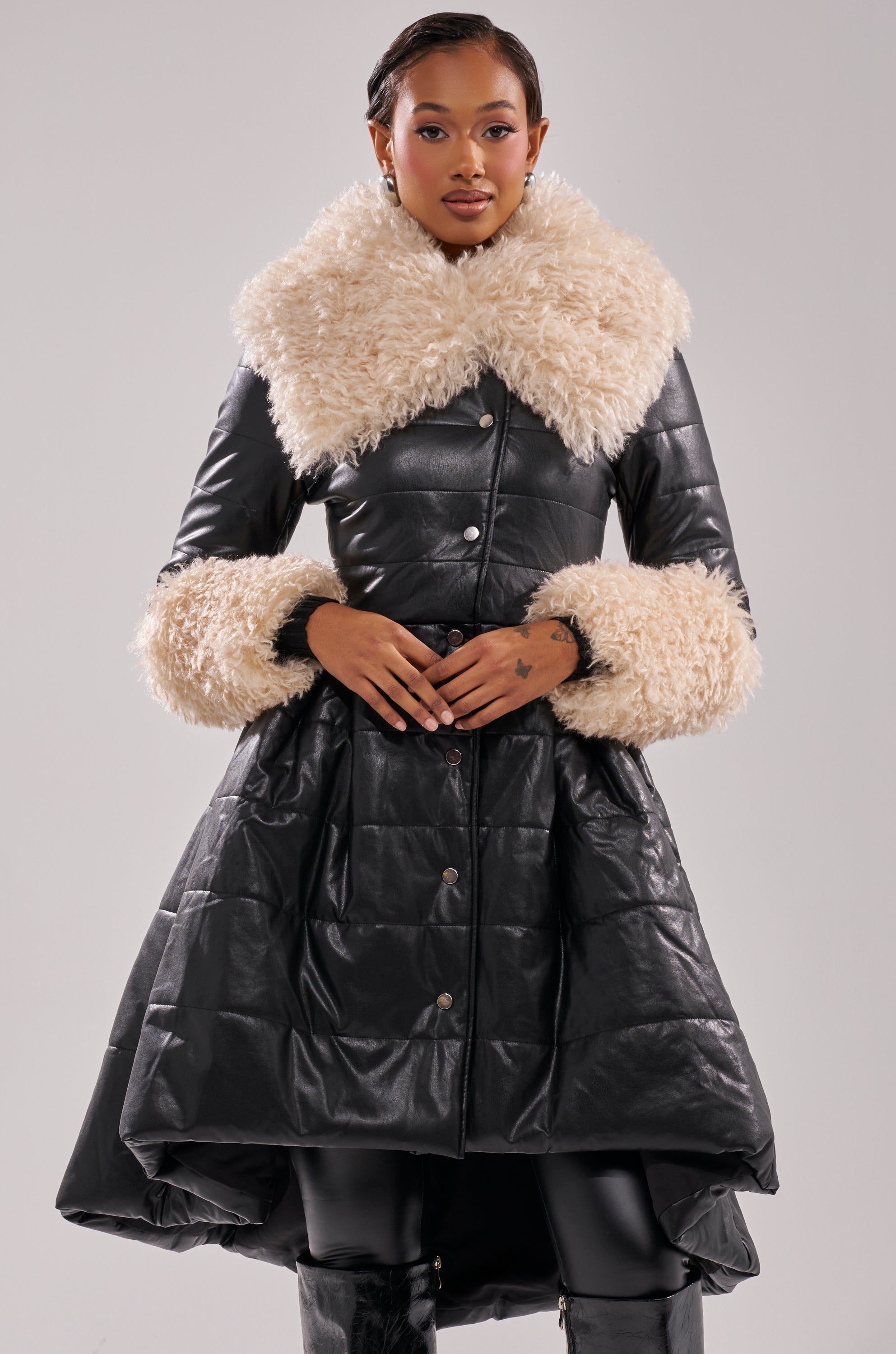 A woman models the MIDTOWN PEPLUM PUFFER IN FAUX LEATHER, featuring a black faux leather exterior and voluminous cream-colored faux fur trim on the collar and cuffs. She stands confidently against a plain light background.