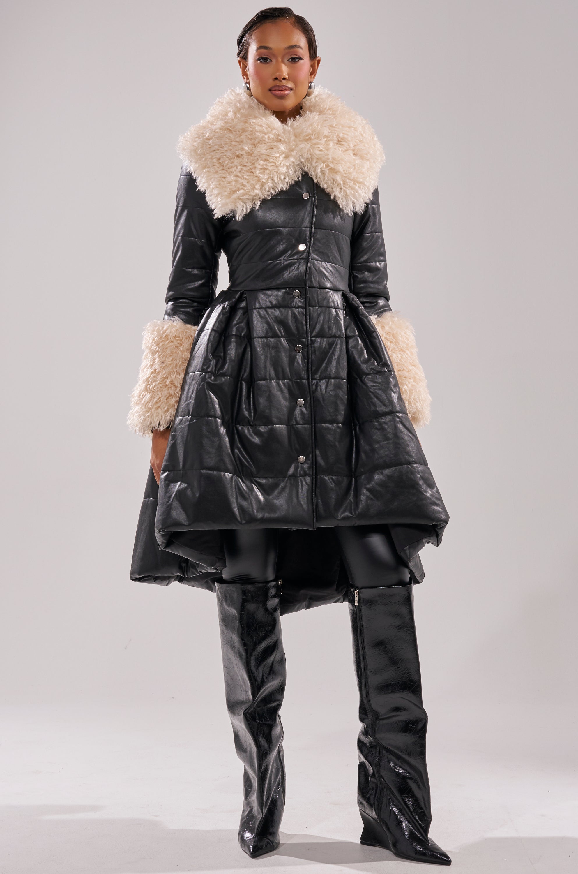 A woman models the Midtown Peplum Puffer in faux leather, a black, knee-length quilted coat with cream faux fur at the collar and cuffs, styled with shiny black thigh-high boots against a plain light gray background.