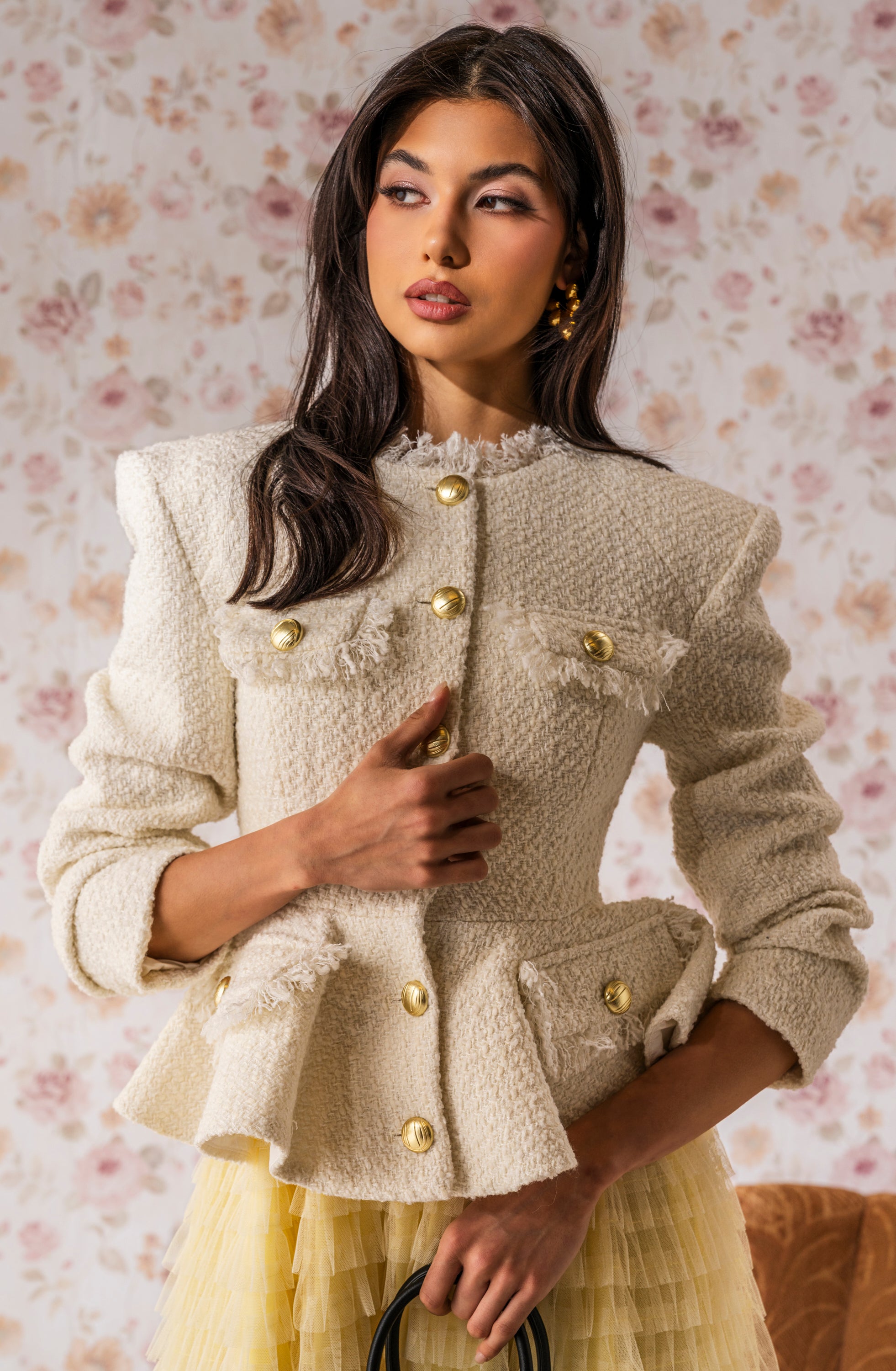 A woman with long dark hair and a confident look stands against floral wallpaper, holding a black handbag while wearing the MALLORCA TWEED BLAZER featuring gold buttons and fringe details. 0