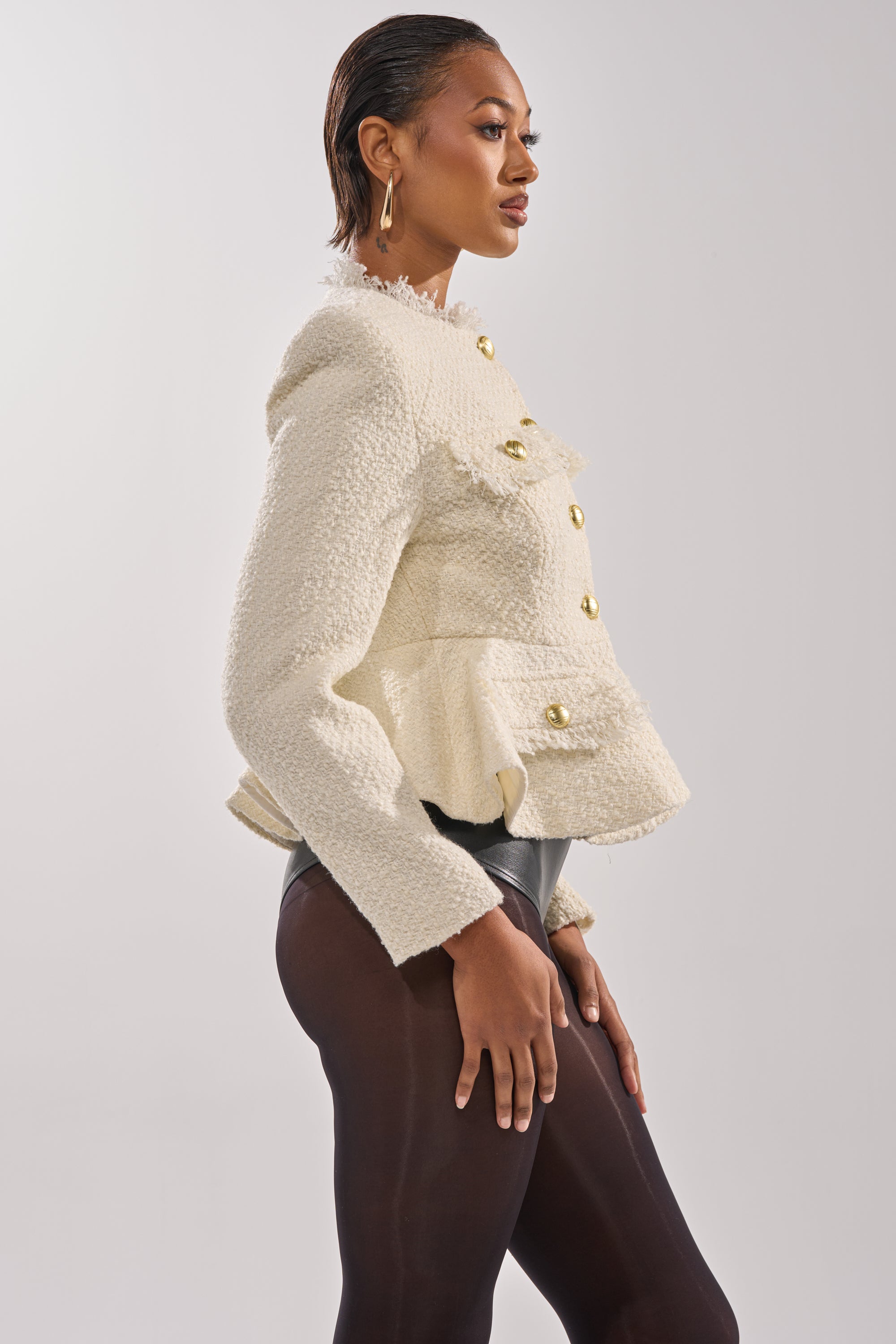 A woman in profile poses confidently in the cream MALLORCA TWEED BLAZER with gold buttons, paired with black tights and gold earrings against a plain light background. 3