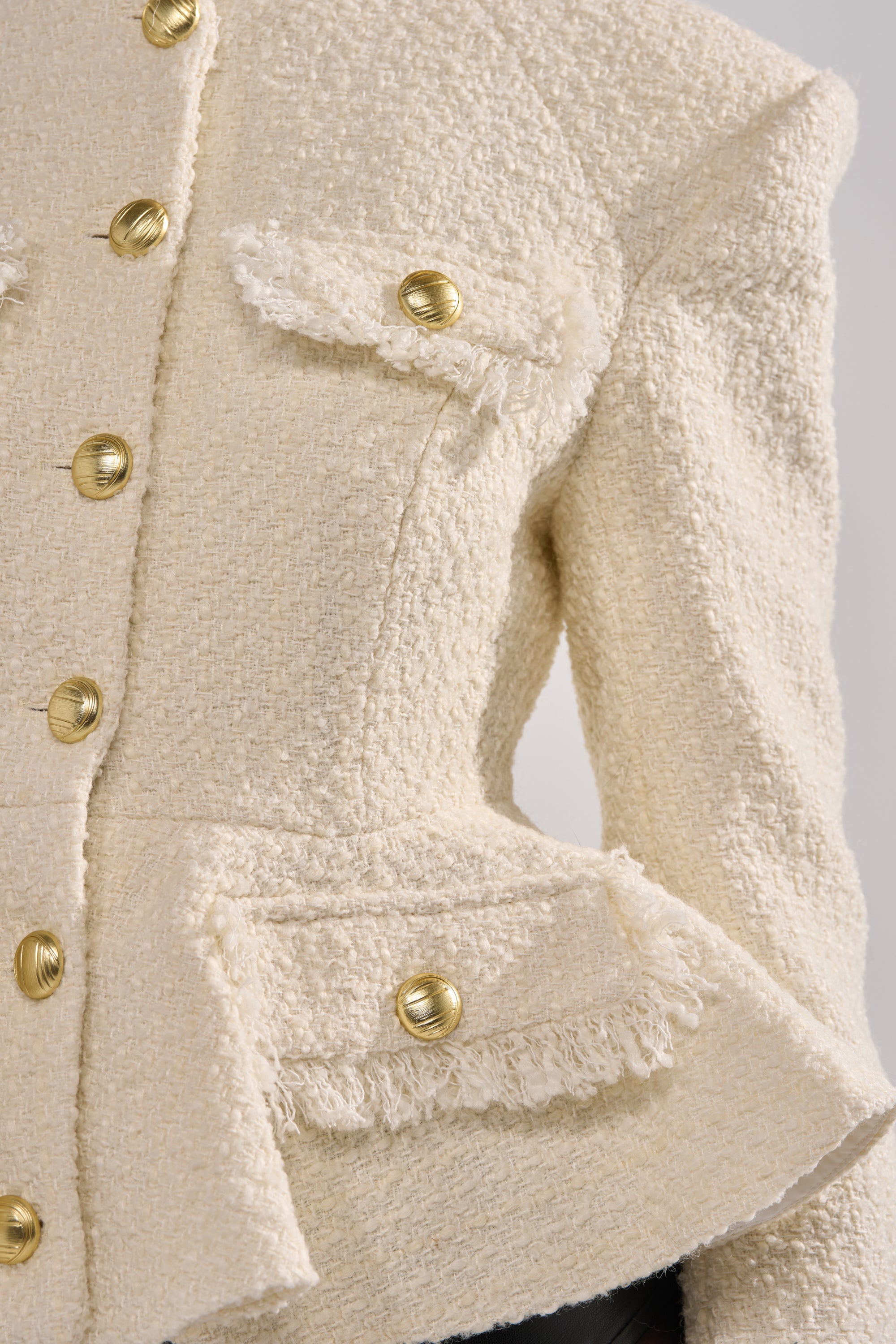 A close-up of the MALLORCA TWEED BLAZER reveals a cream textured tweed blazer featuring gold buttons, flap pockets with fringe trim, and standout button details for a structured, elegant look. 7