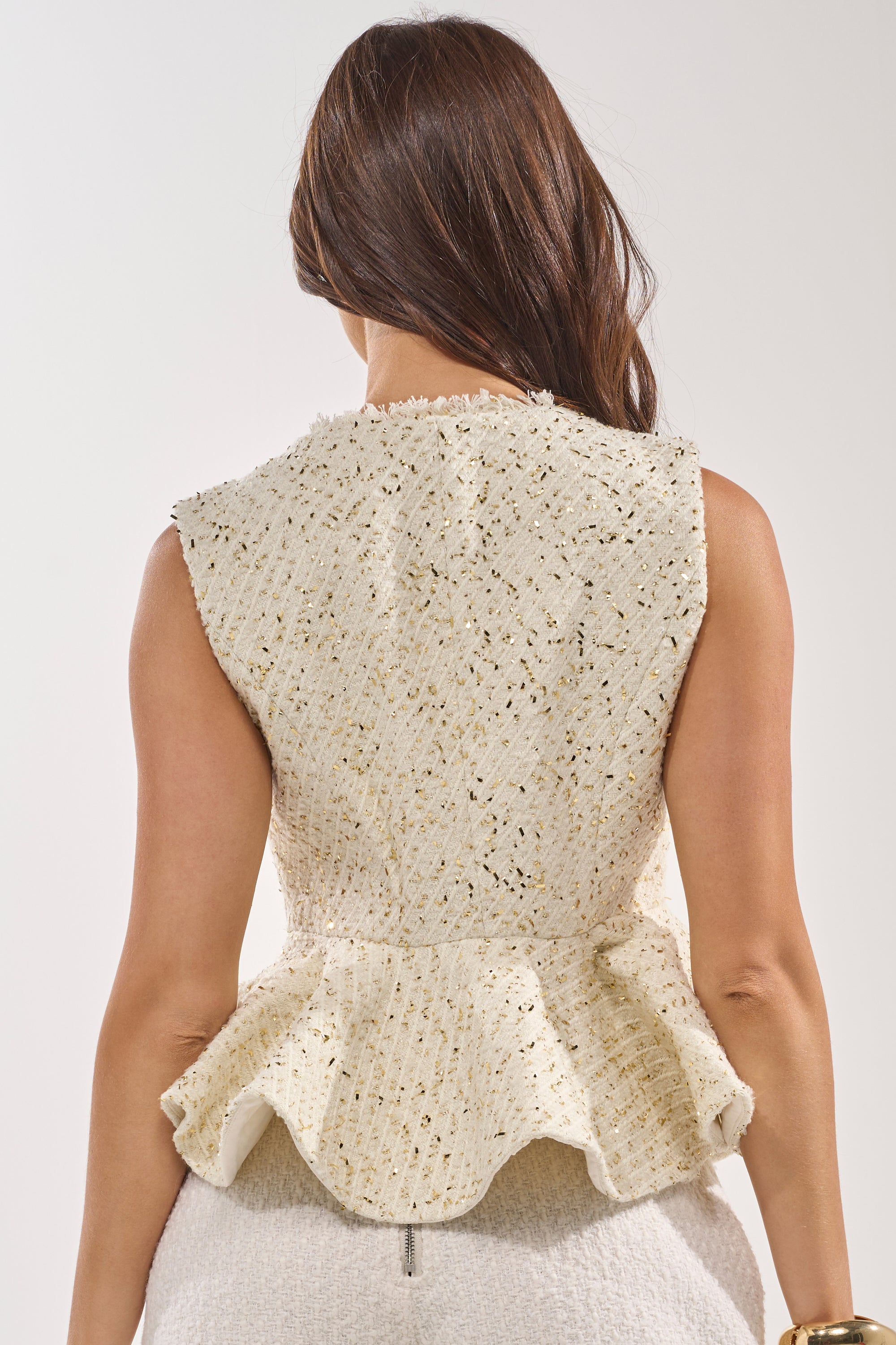 A woman with long brown hair, seen from behind, wears the MALLORCA TWEED VEST in beige featuring a structured sleeveless design, ruffled peplum hem, matching bottoms, and subtle metallic gold accents. 3