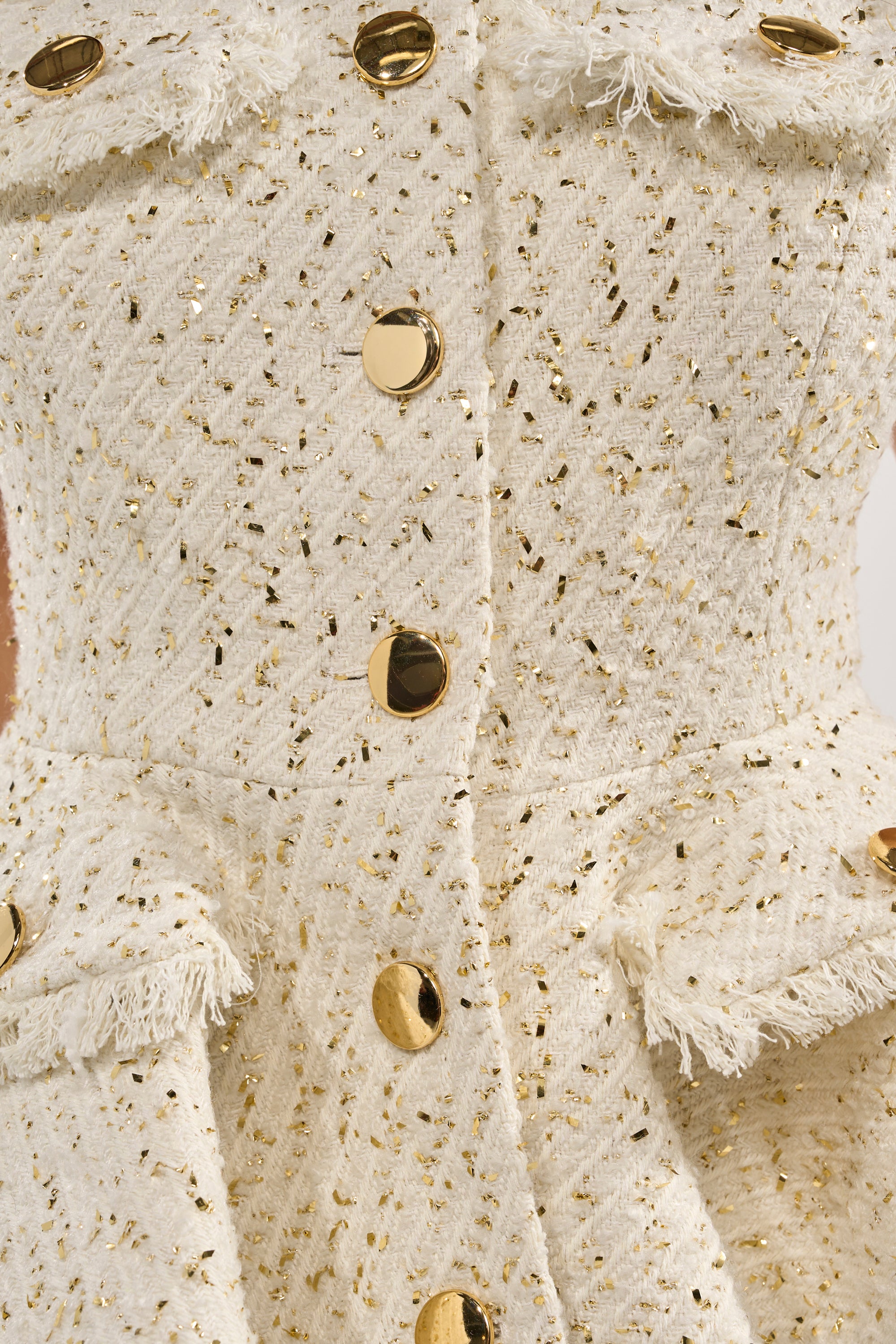 Close-up of the MALLORCA TWEED VEST: a cream-colored textured fabric vest with metallic gold detailing, shiny gold buttons, and two fringed flap pockets for a refined, structured sleeveless look. 6