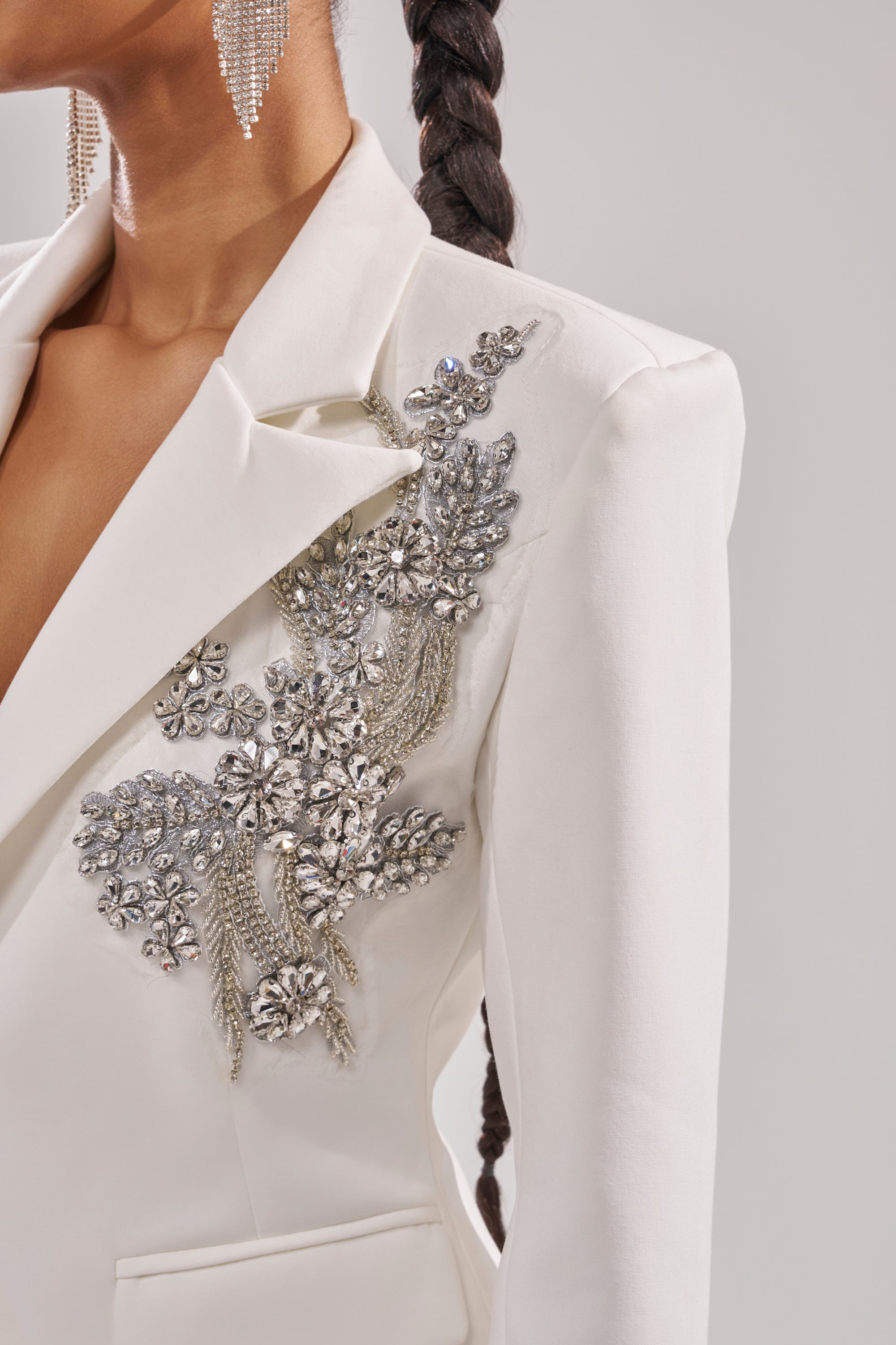A woman styles the SPEND IT ALL ON ME EMBELLISHED BLAZER, featuring intricate silver crystal lapel details, with dangling rhinestone earrings and a long braided ponytail. 9