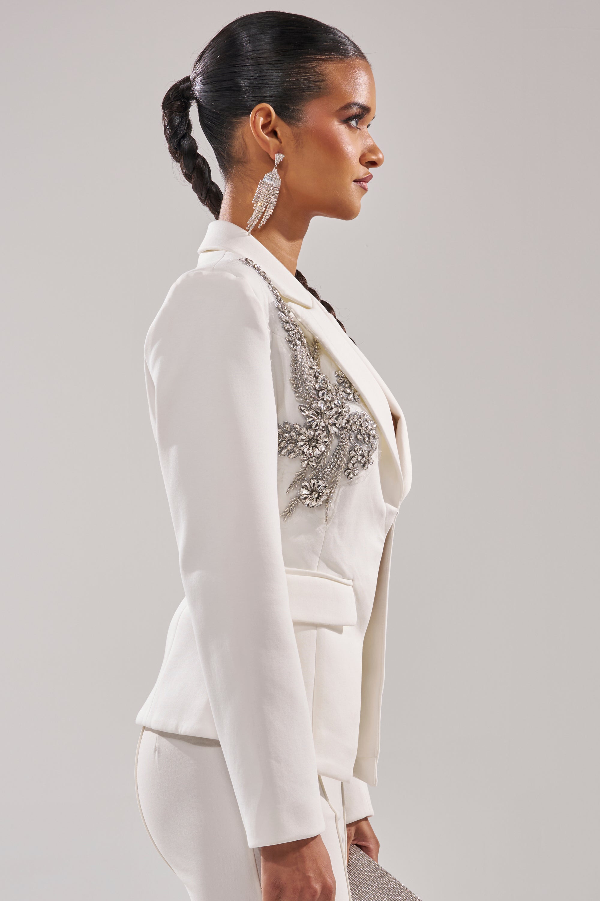 A woman in profile with a sleek braided ponytail models the SPEND IT ALL ON ME EMBELLISHED BLAZER, featuring intricate silver detailing, paired with large dangling earrings against a plain background. 4