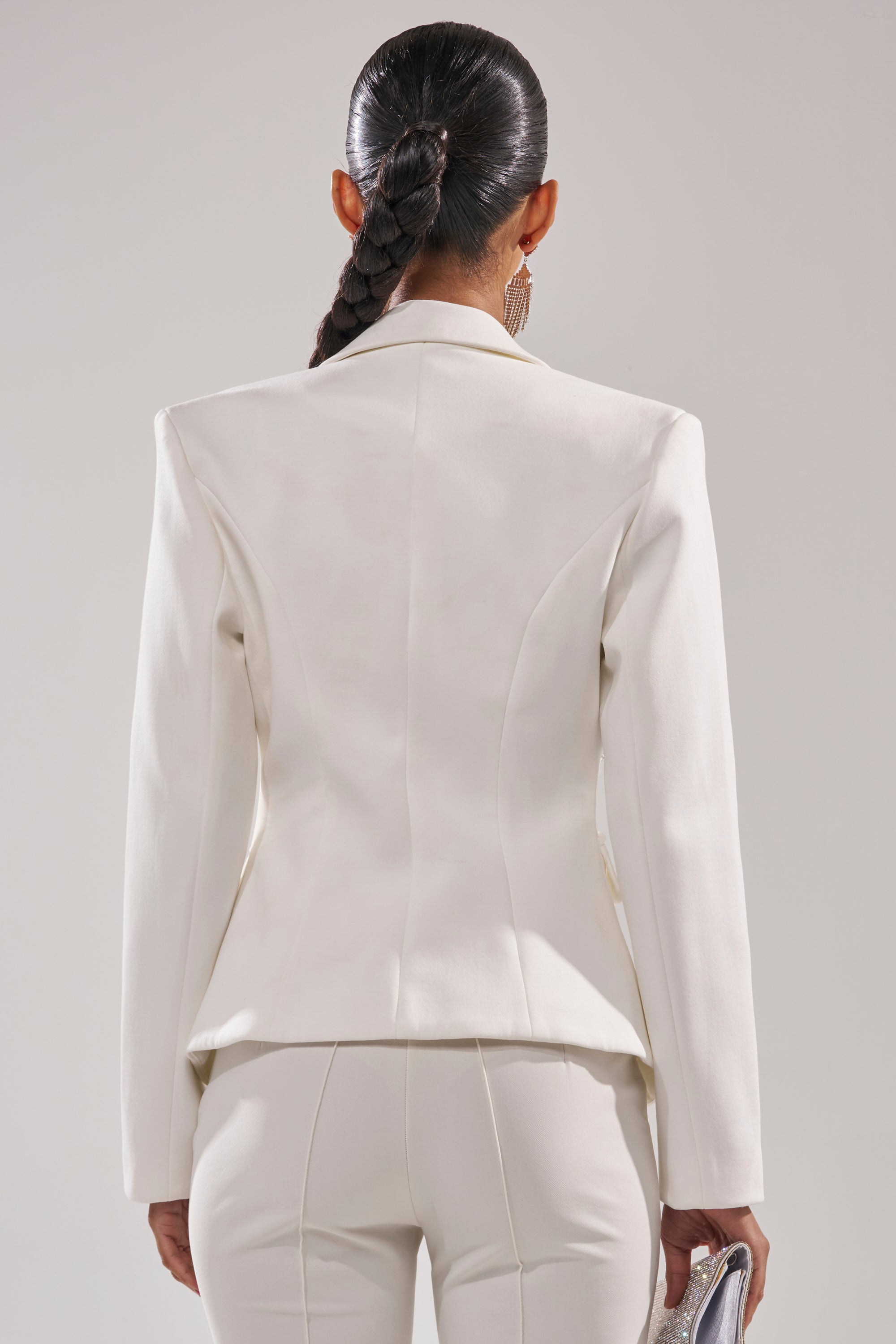 A woman with a long braided ponytail is shown from the back wearing the SPEND IT ALL ON ME EMBELLISHED BLAZER and matching pants, accessorized with statement earrings and holding a metallic clutch in her left hand. 5