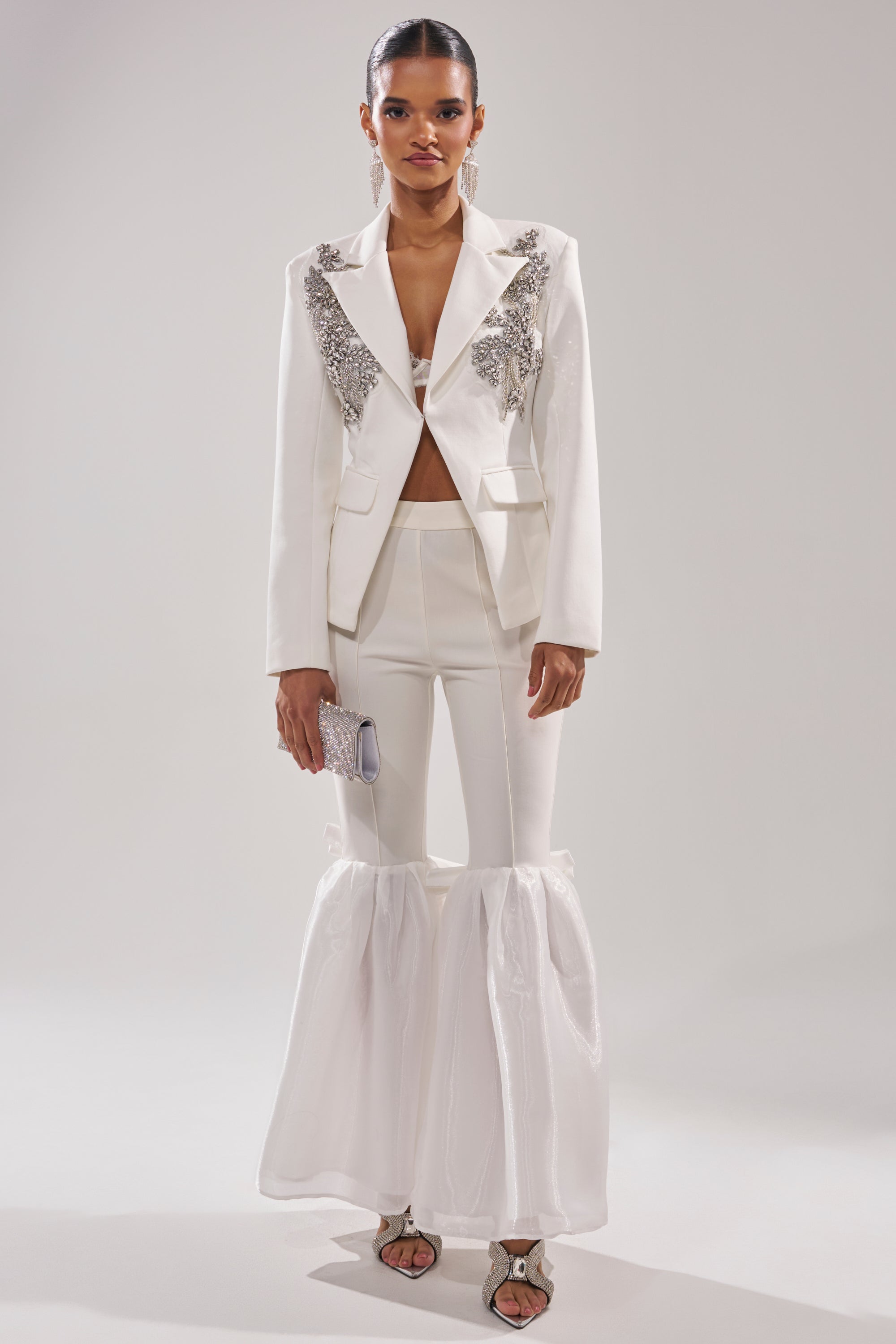 A model wears the SPEND IT ALL ON ME EMBELLISHED BLAZER in white with silver accents, paired with flared sheer-bottom fitted pants, open-toe heels, and a small silver clutch against a plain background. 6