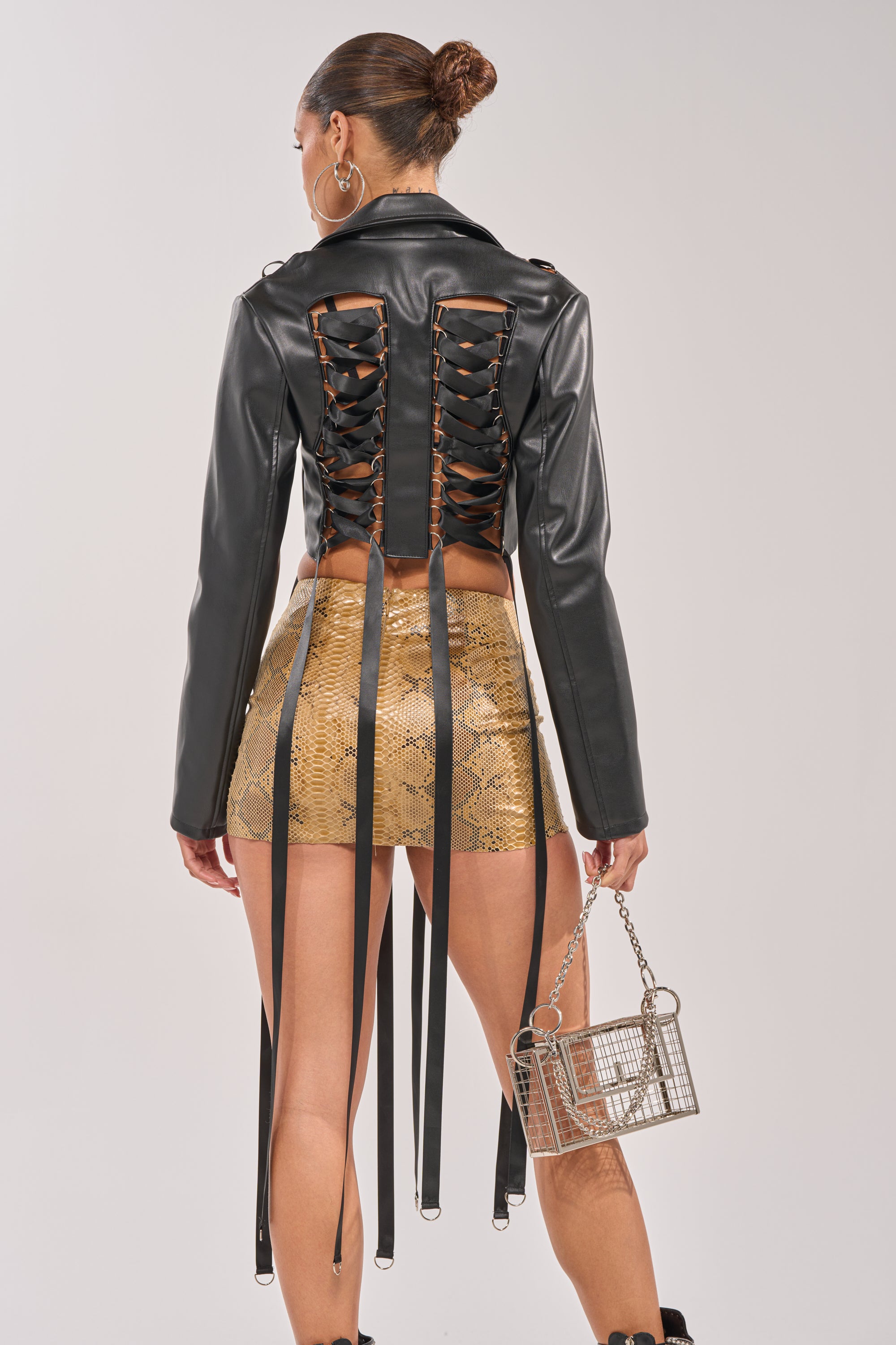 A woman stands facing away, wearing the IN THE MOMENT LACE UP BLAZER in black faux leather with lace-up details and long straps, paired with a gold snakeskin-patterned mini skirt and a metallic cage handbag. 4