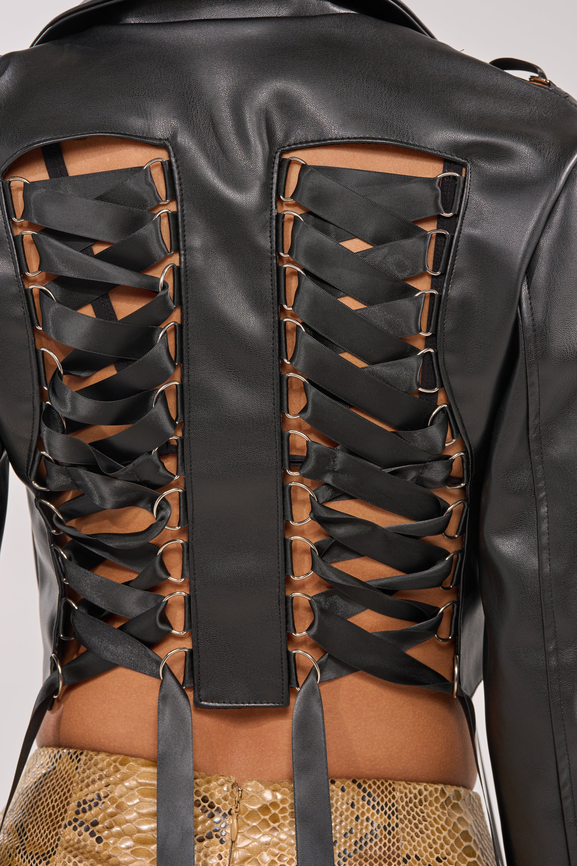 A close-up of someone wearing the IN THE MOMENT LACE UP BLAZER, featuring wide black ribbon laces crisscrossing over two open back panels, paired with tan snake-print textured pants. 8