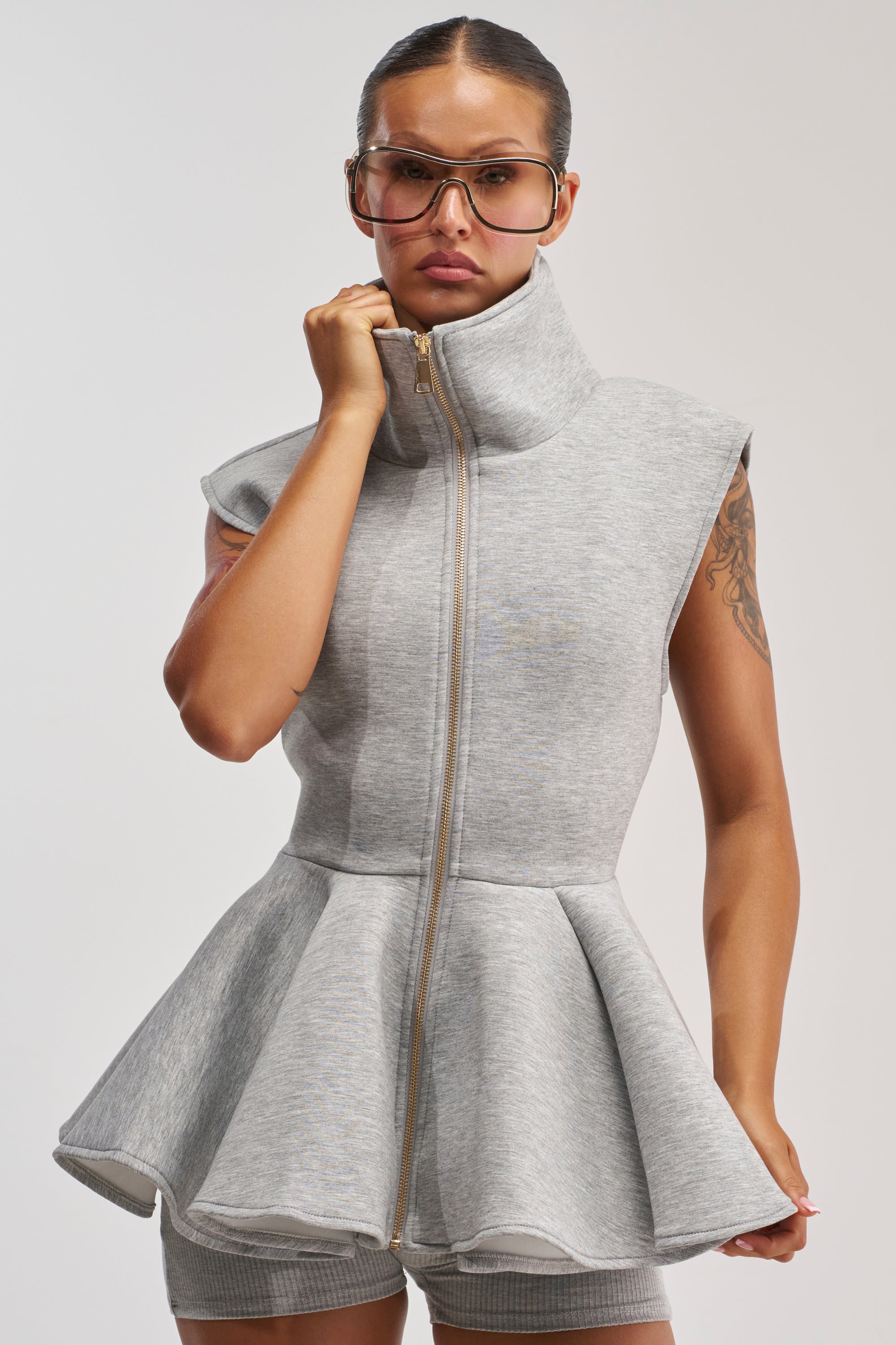 A woman wears the LOTTIE NEOPRENE PEPLUM VEST IN HEATHER GREY—sleeveless with a tailored, waist-defining peplum silhouette—paired with matching shorts and large clear glasses, her hand holding the zipper against a plain background.