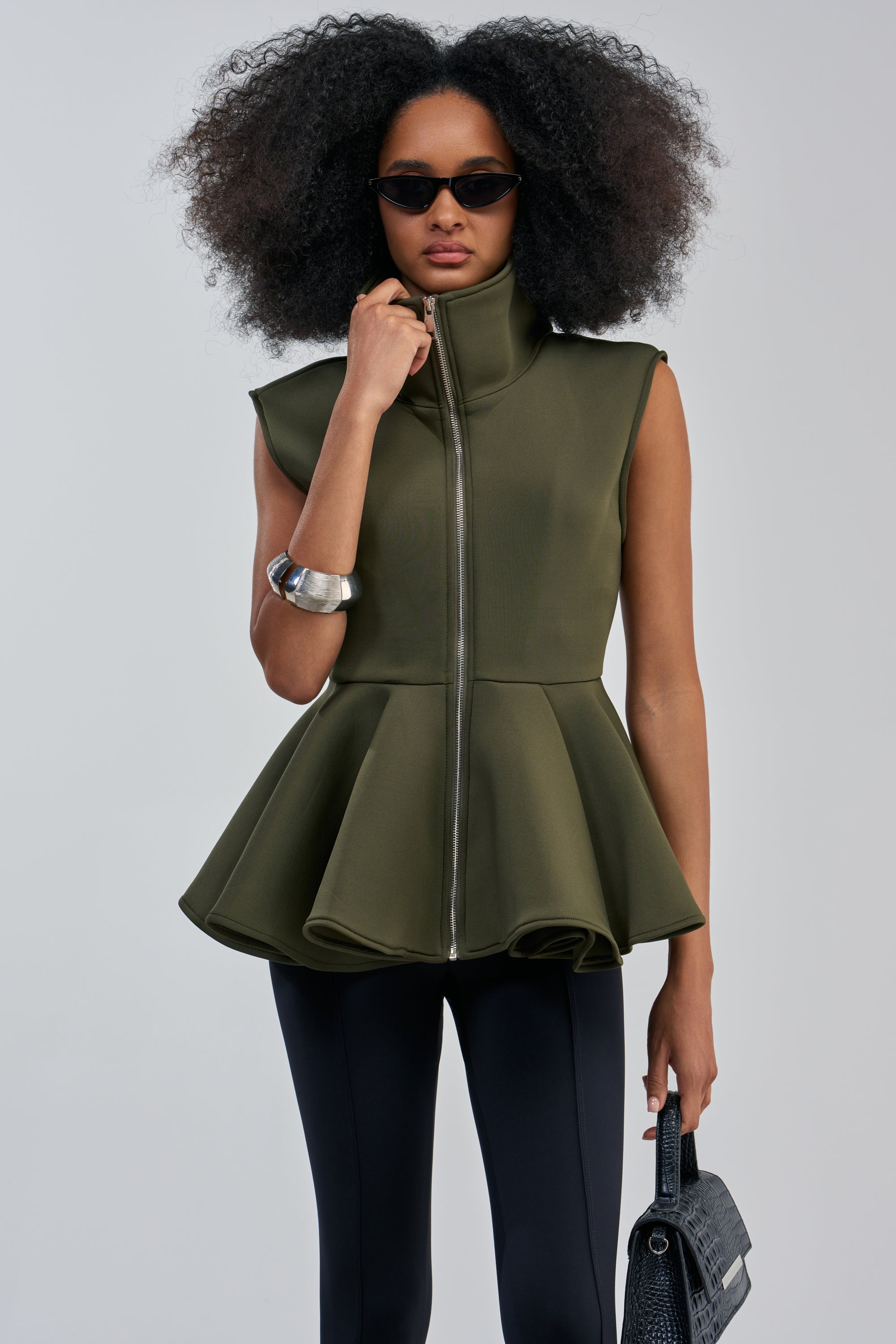 A woman in sunglasses wears the LOTTIE NEOPRENE PEPLUM VEST IN OLIVE with black leggings and carries a textured black handbag. Her voluminous curly hair and silver cuff bracelet complete an effortlessly chic look, perfect for fall layering.