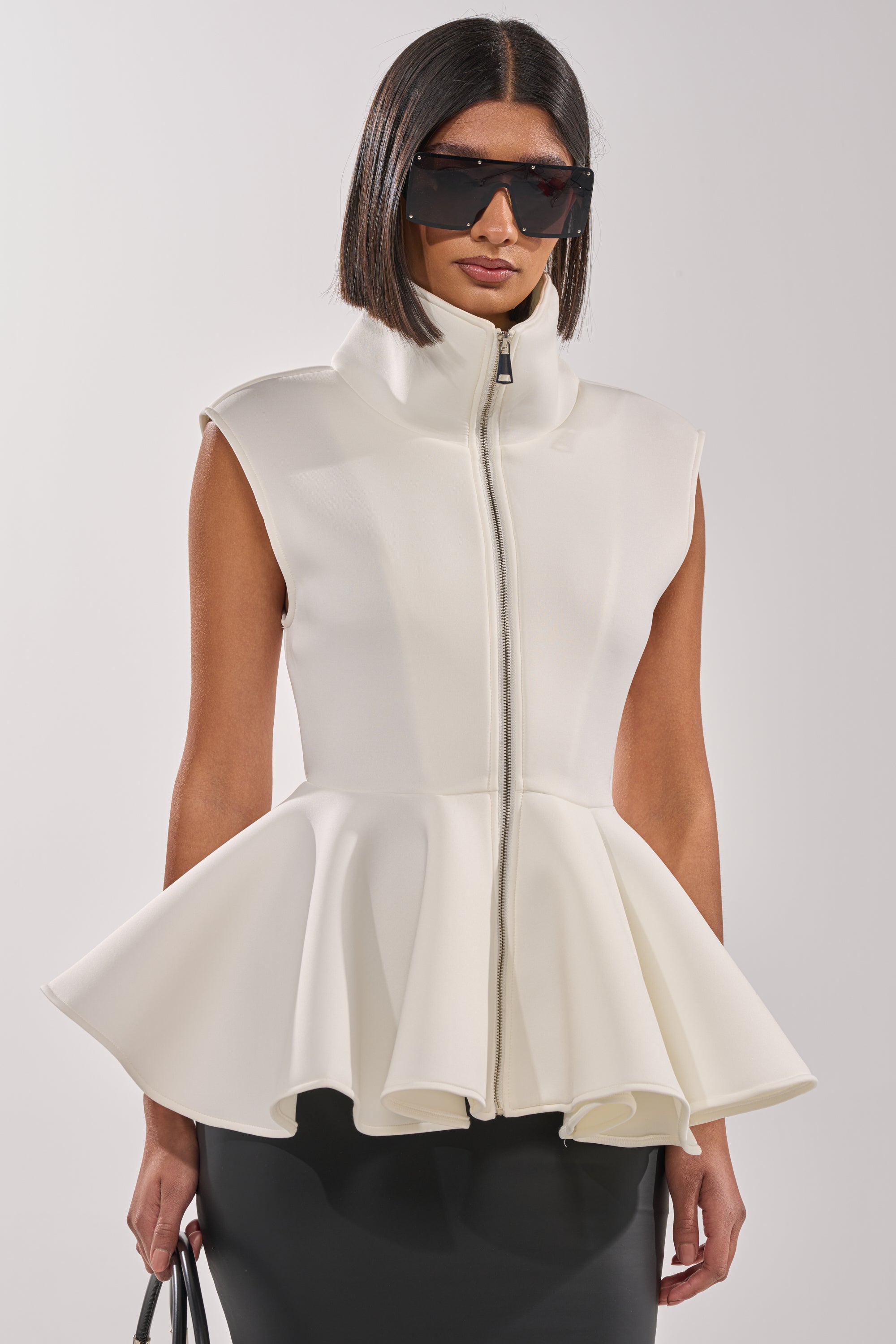 A woman styles the LOTTIE NEOPRENE PEPLUM VEST—an ivory sleeveless top with a high collar and flared peplum hem—with large black sunglasses and a black handbag for a chic look. 0