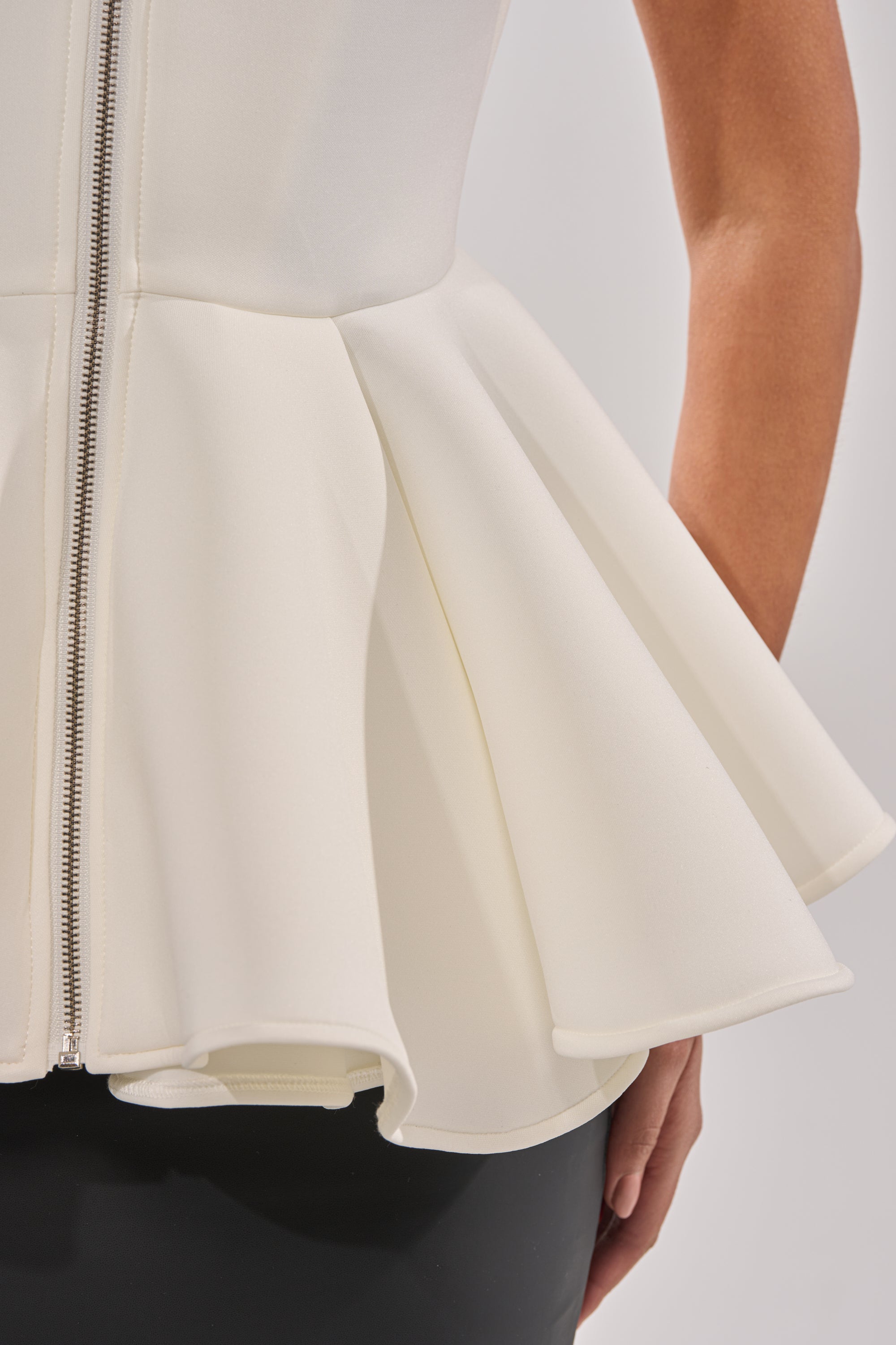 Close-up of a person wearing the LOTTIE NEOPRENE PEPLUM VEST, a sleeveless ivory collared top with visible zipper and structured, flared waist. The person's arm and part of a black garment are also seen. 9