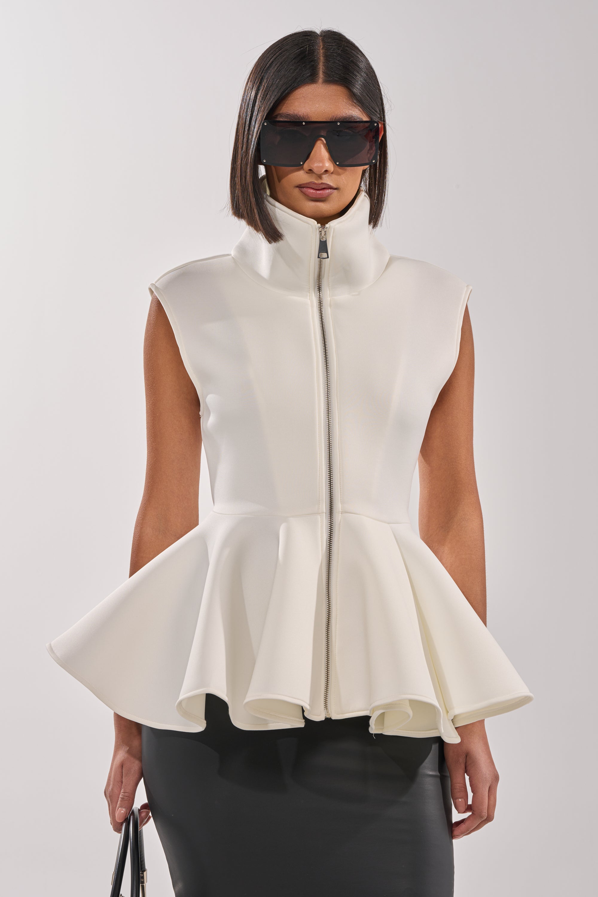 A woman in large black sunglasses and the LOTTIE NEOPRENE PEPLUM VEST, paired with a black skirt and black handbag, stands against a plain light background. 1
