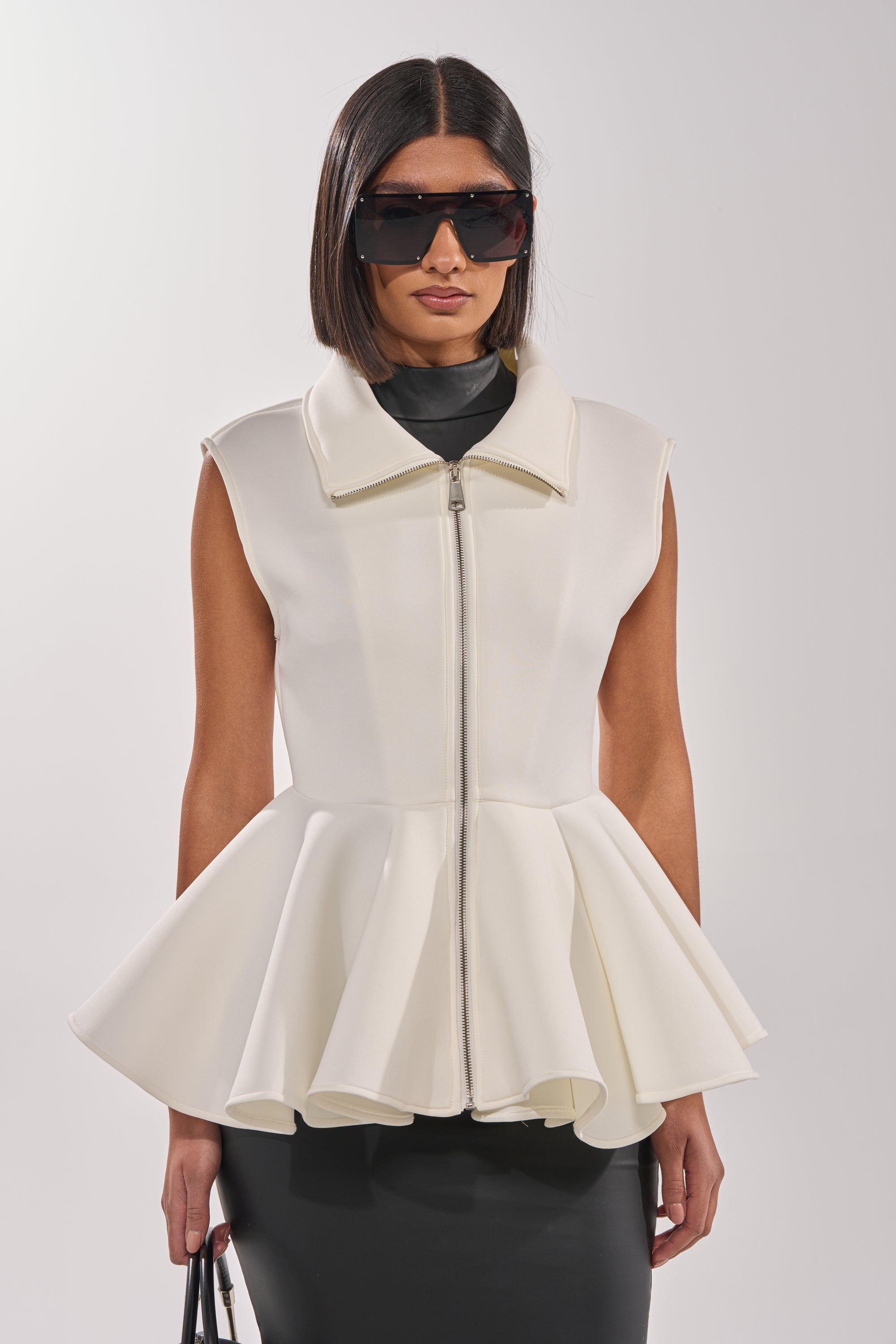 A woman with a sleek bob wears oversized sunglasses and the LOTTIE NEOPRENE PEPLUM VEST, holding a black handbag against a plain light background. 2