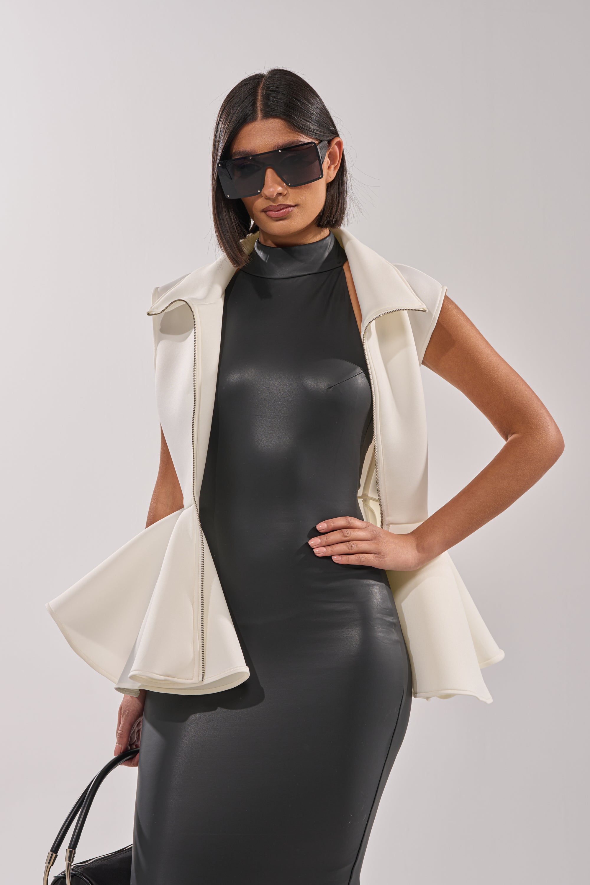 A woman wears the LOTTIE NEOPRENE PEPLUM VEST as a stylish ivory layer over a sleek black sleeveless dress. She accessorizes with large black sunglasses, straight dark hair, and a black handbag against a light gray background. 3