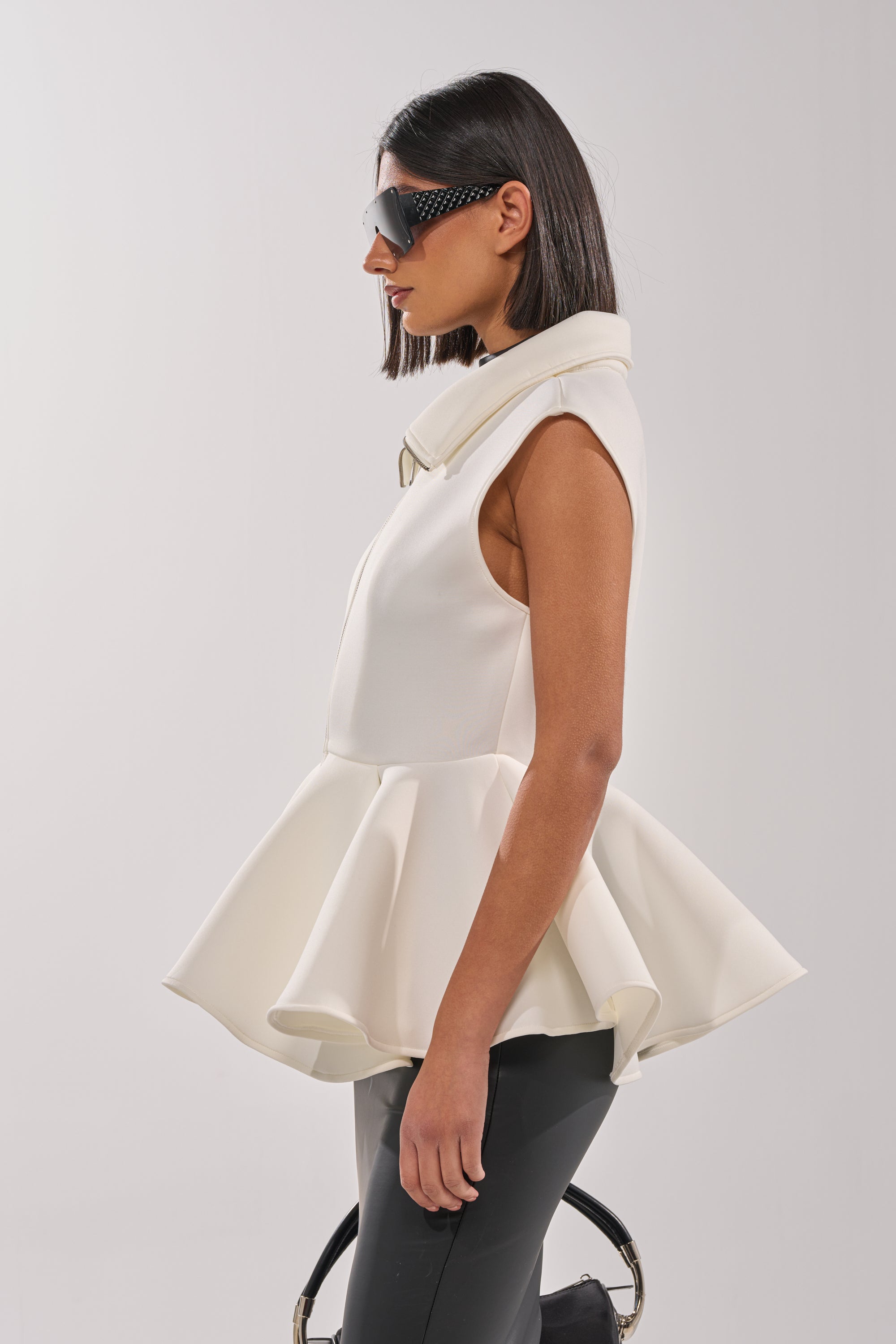 A woman with straight dark hair and large sunglasses models the LOTTIE NEOPRENE PEPLUM VEST, styled with black pants and a bag, standing in profile against a plain background. 5