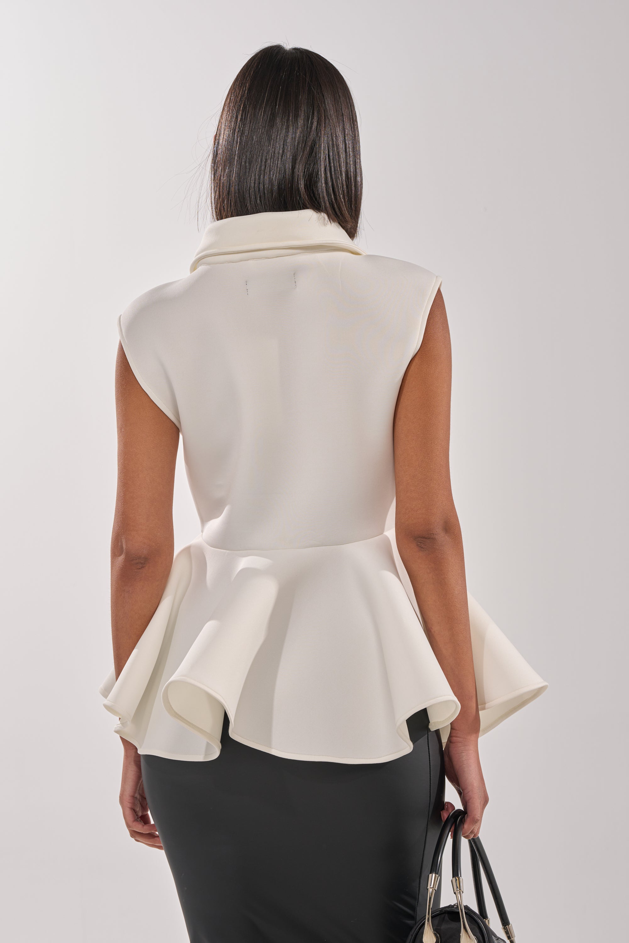 A woman seen from behind wears the LOTTIE NEOPRENE PEPLUM VEST in ivory with a fitted black skirt and holds a black handbag. The background is plain and light-colored. 6