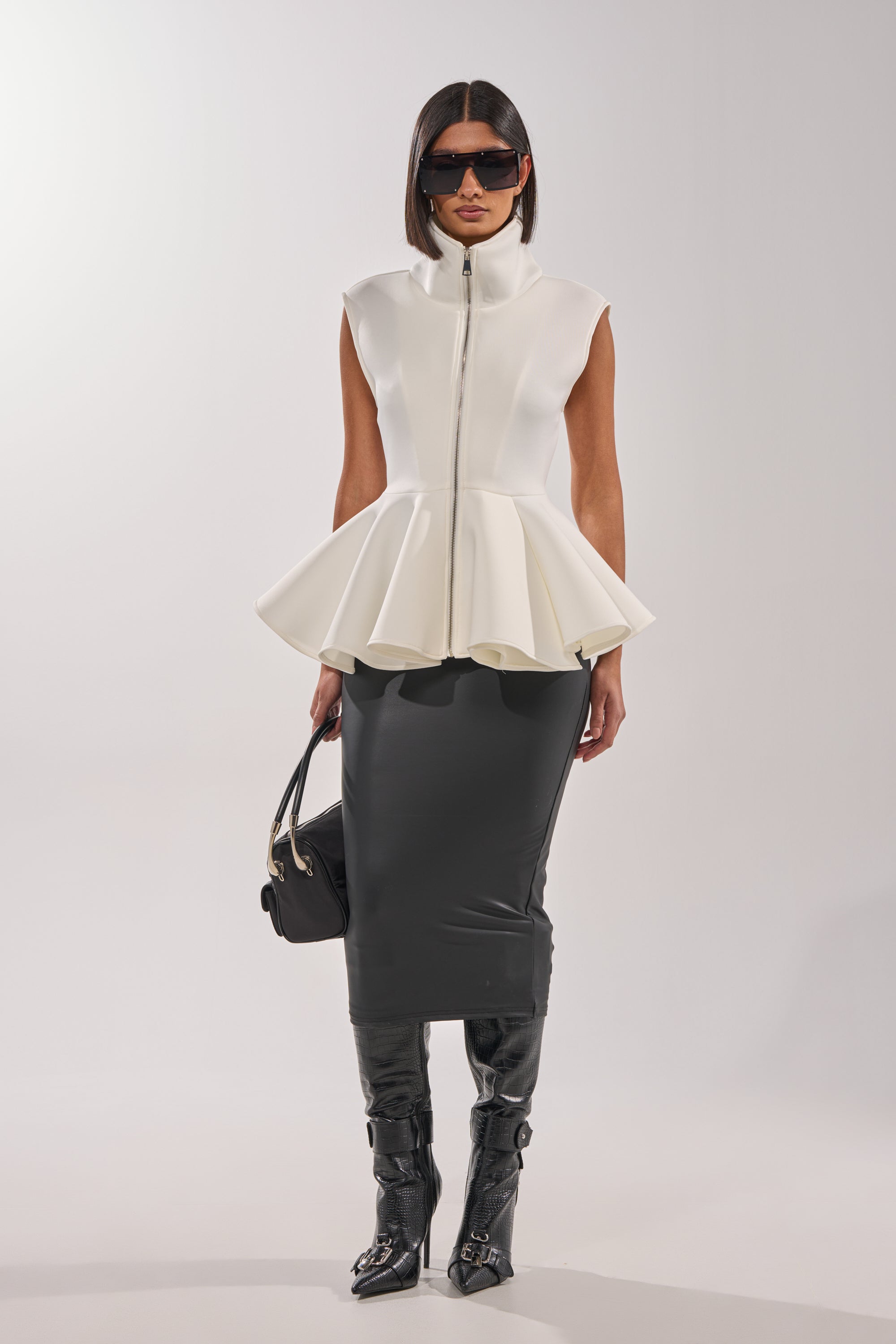 A woman in the LOTTIE NEOPRENE PEPLUM VEST, a fitted black midi skirt, tall black boots, sunglasses, and carrying a black handbag stands against a plain light background. 7