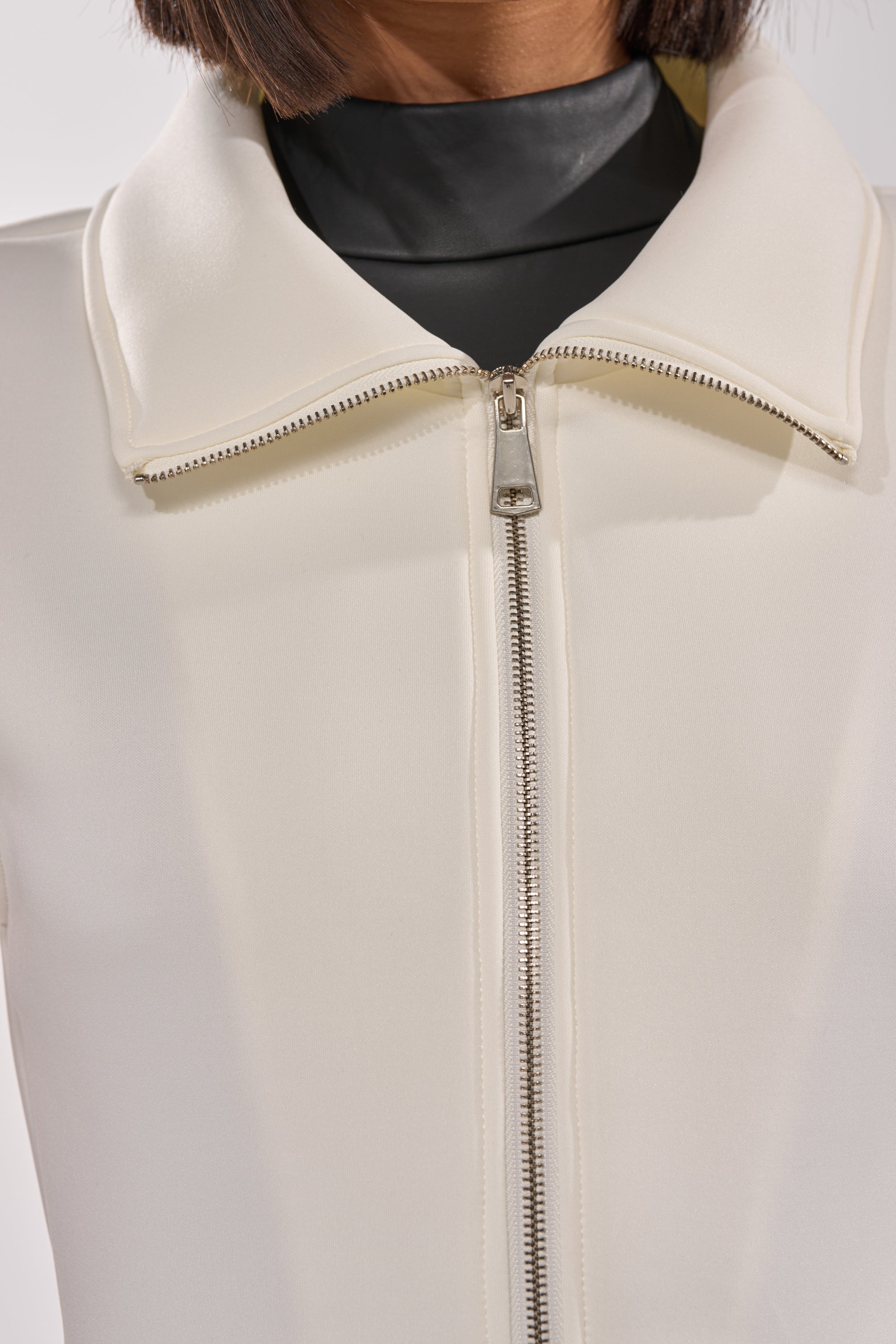 Close-up of the LOTTIE NEOPRENE PEPLUM VEST in ivory, featuring a wide collar and zipper, layered over a black high-neck top. The focus is on the vest’s zipper and upper section; the wearer’s face is not shown. 8