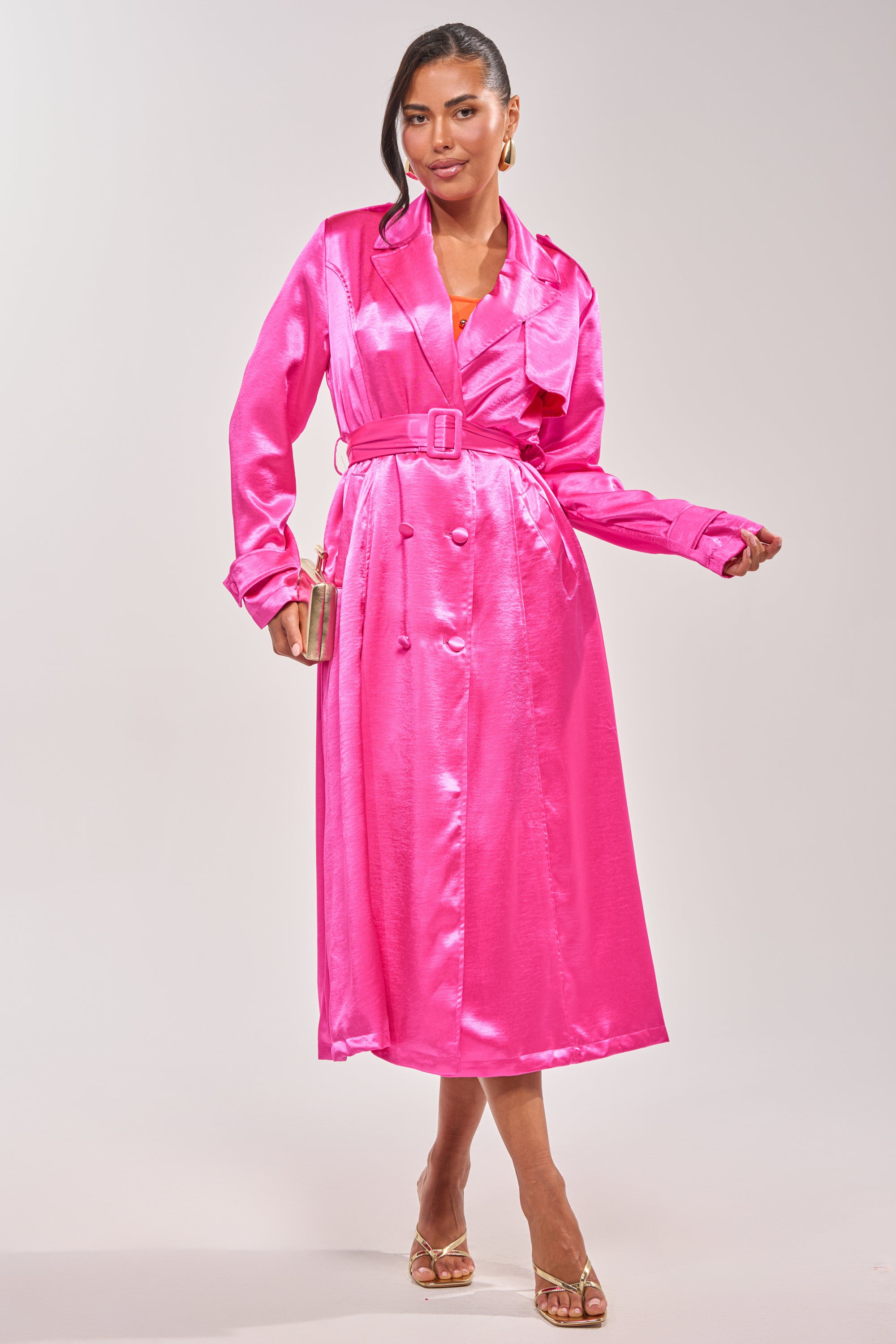Wearing the FOR THE DRAMA SATIN TRENCH in hot pink with a belt and gold heels, she poses against a plain light backdrop holding a metallic clutch—a bold statement piece for date night. 0