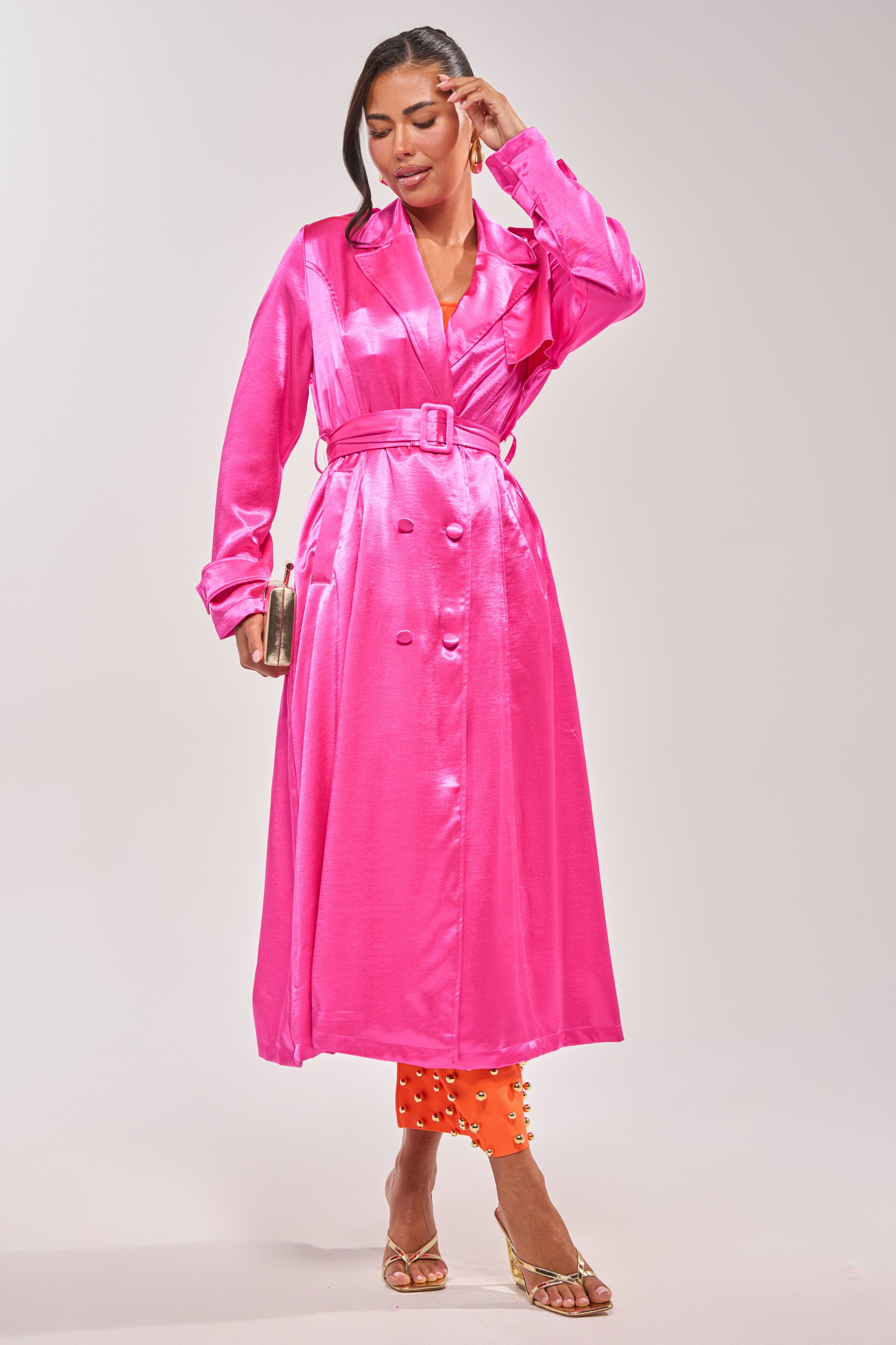 A woman in the FOR THE DRAMA SATIN TRENCH, styled over orange embellished pants, with a clutch and strappy heels—statement layering at its finest against a plain backdrop. 1