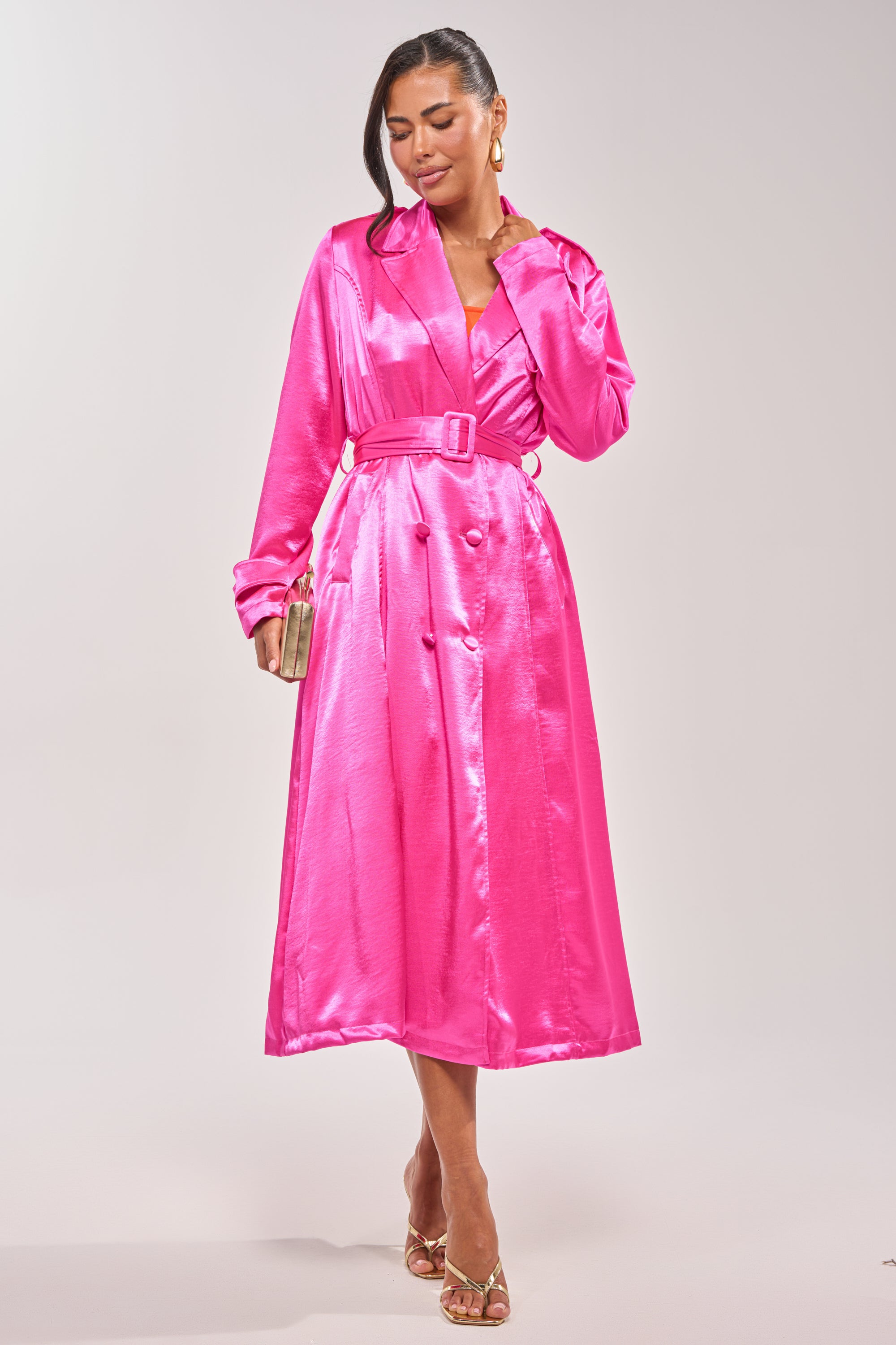 Wearing the FOR THE DRAMA SATIN TRENCH in hot pink with gold hoop earrings, tan heels, and a gold clutch, she poses against a plain background, smiling with her hand touching her neck—ideal for statement layering or a date night. 2