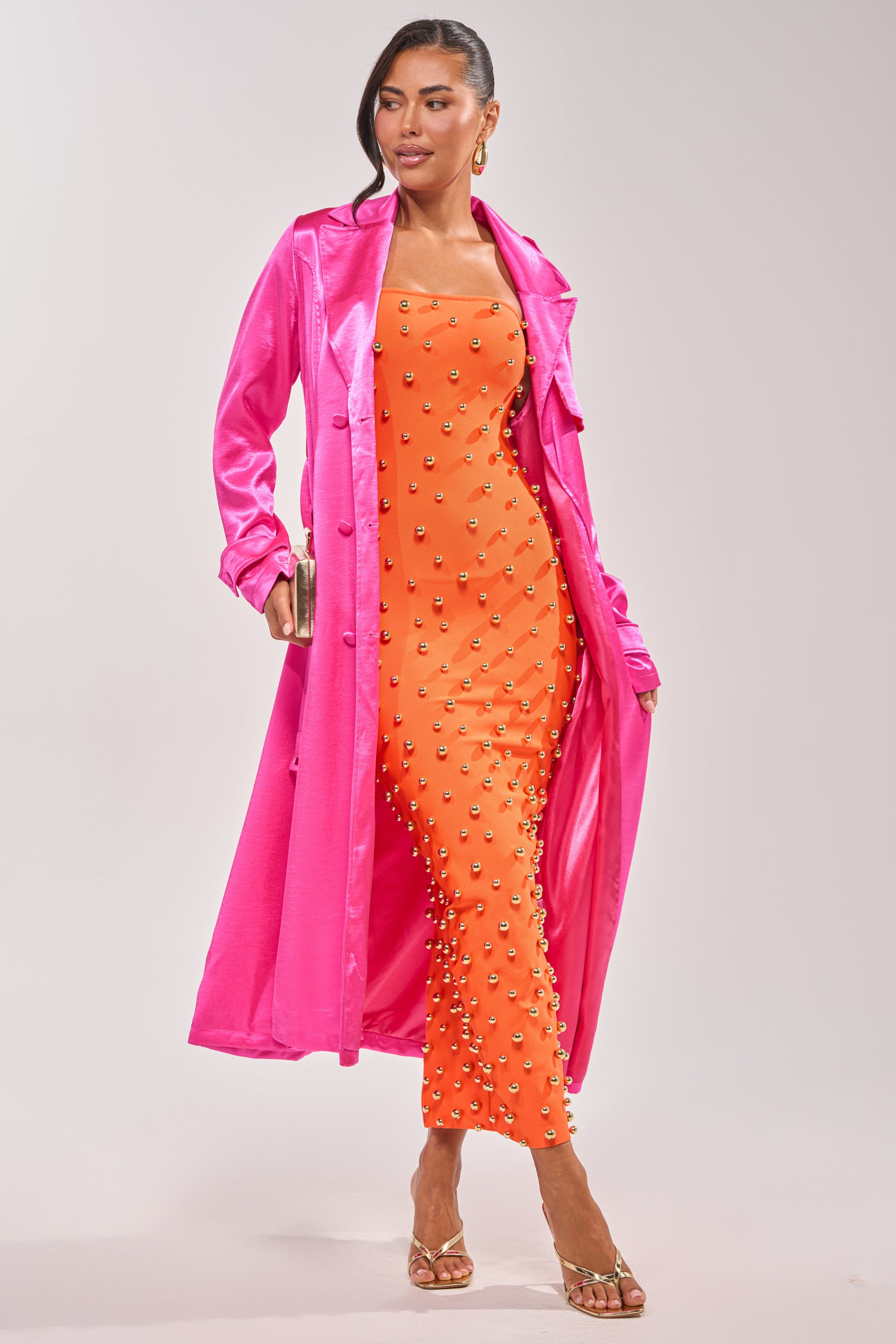 A woman wears the FOR THE DRAMA SATIN TRENCH in hot pink over a bright orange pearl-embellished dress, styled with a gold clutch and sandals—an eye-catching look for date night. 4