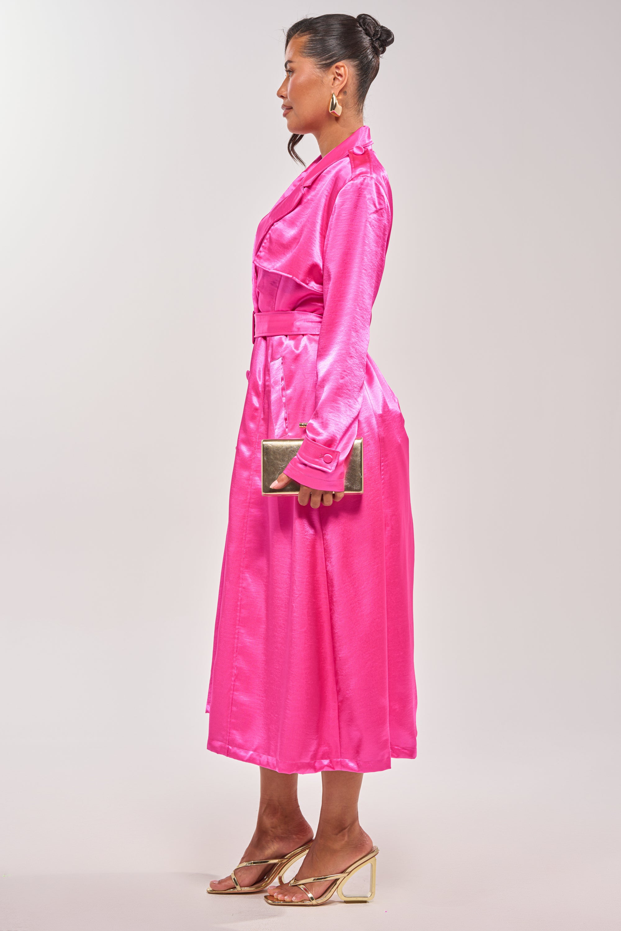 A woman in profile wears the FOR THE DRAMA SATIN TRENCH, a hot pink midi dress with long sleeves. She styles her hair in a neat bun, accessorizes with gold hoop earrings, a gold clutch, and clear high-heeled sandals. 5